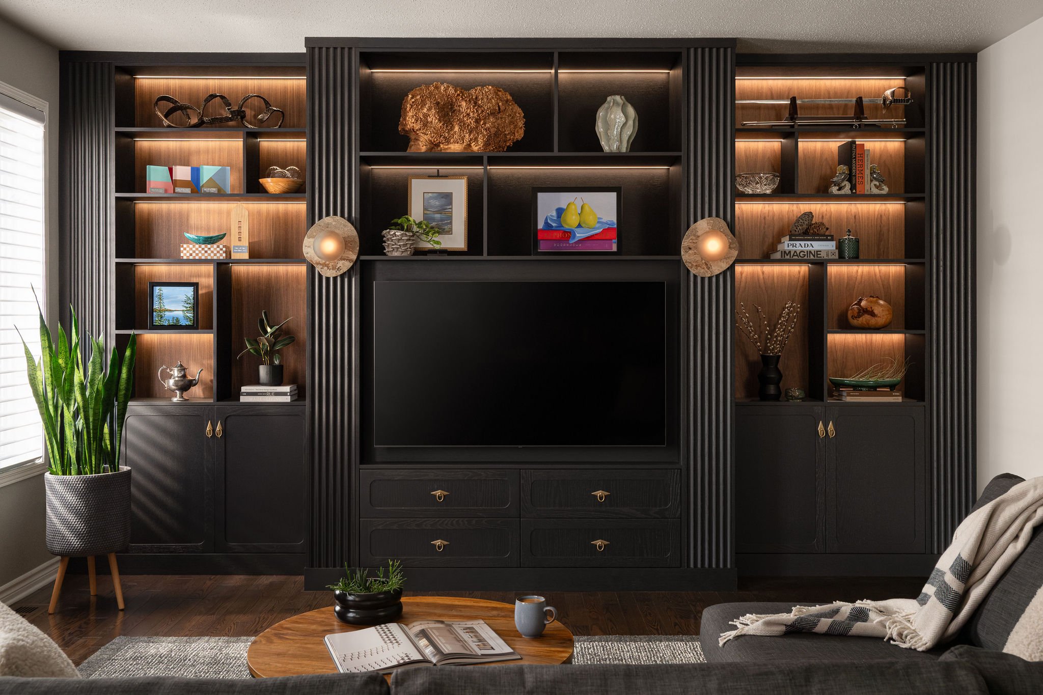 Custom black media wall with fluted details, warm wood open shelving, integrated lighting, brass hardware, large television, styled decor, and grey sectional sofa.