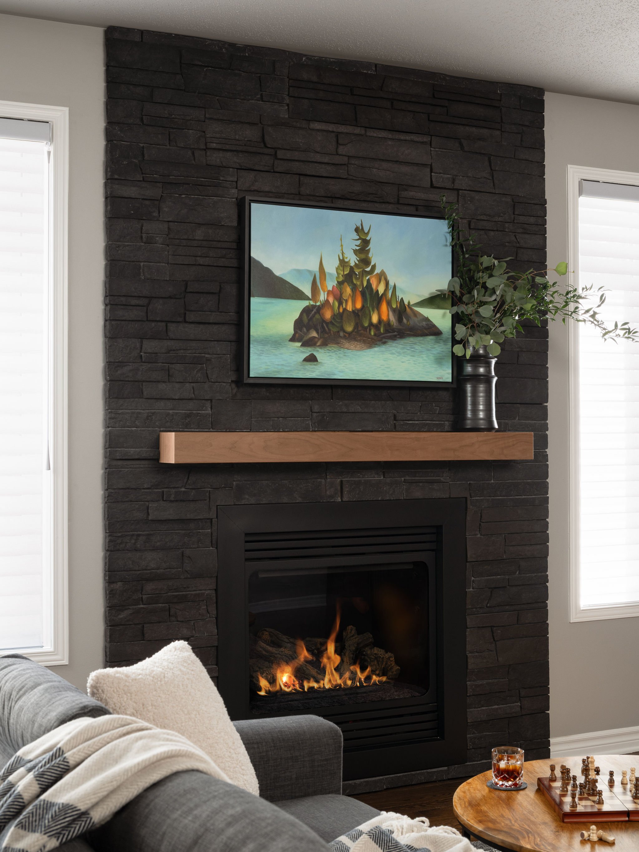 Dark stone fireplace with wood mantel, framed landscape artwork, gas fire, grey sofa, round wood coffee table, and chess board.