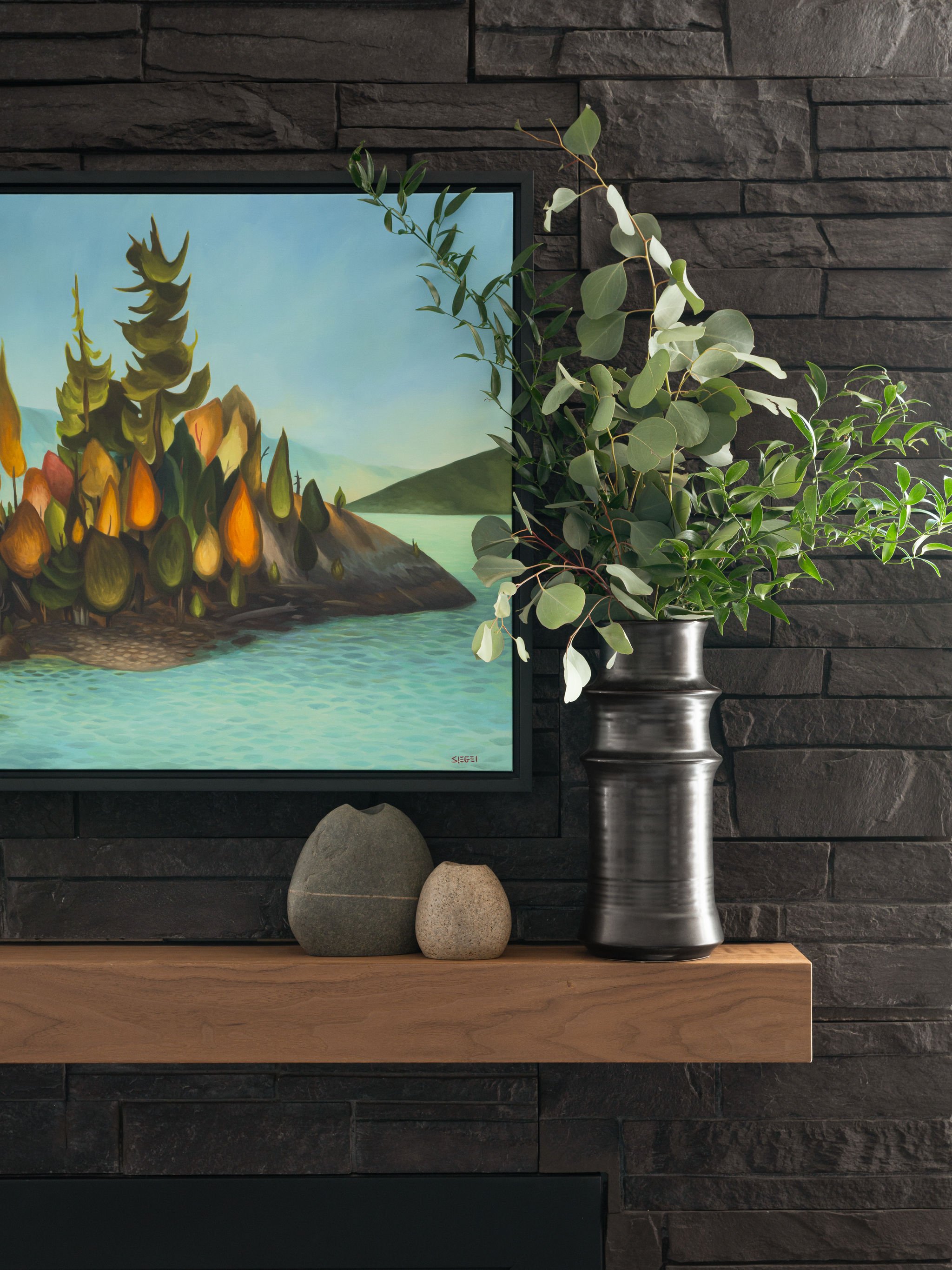 Close-up of dark stone fireplace mantel styled with Canadian landscape artwork, black vase with greenery, and natural stone decorative objects.