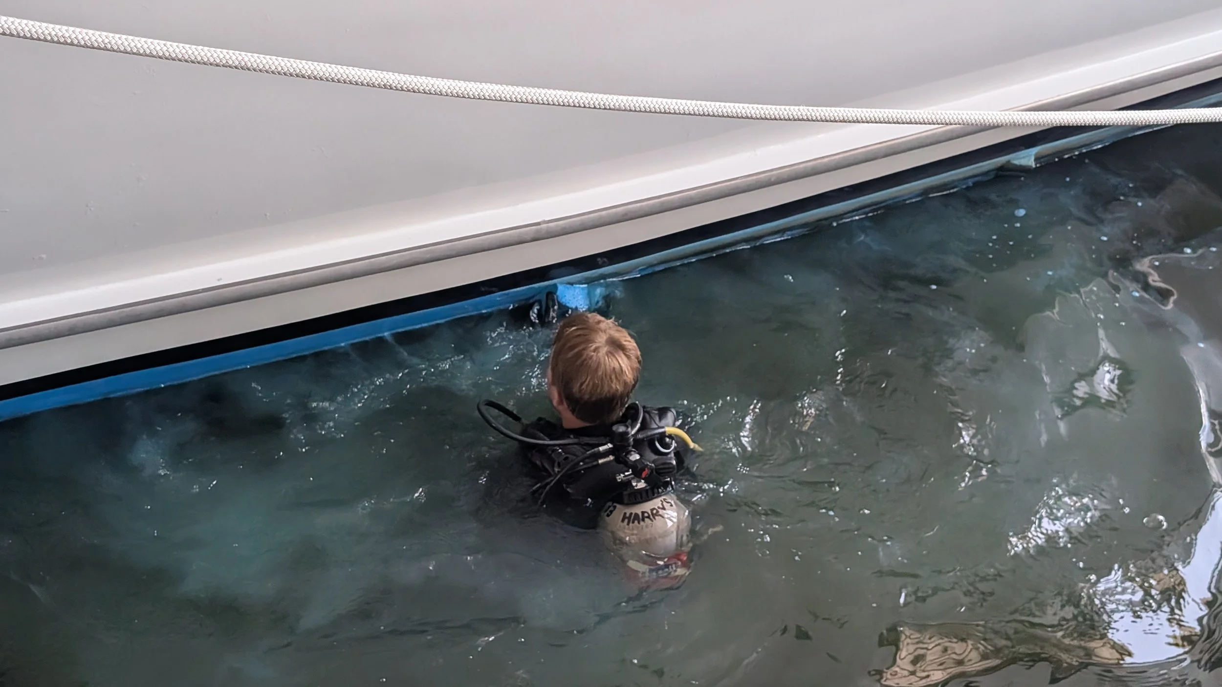 A person in scuba gear is submerged in water next to a boat, with their back to the camera, wearing a helmet labeled 'HARRY'.