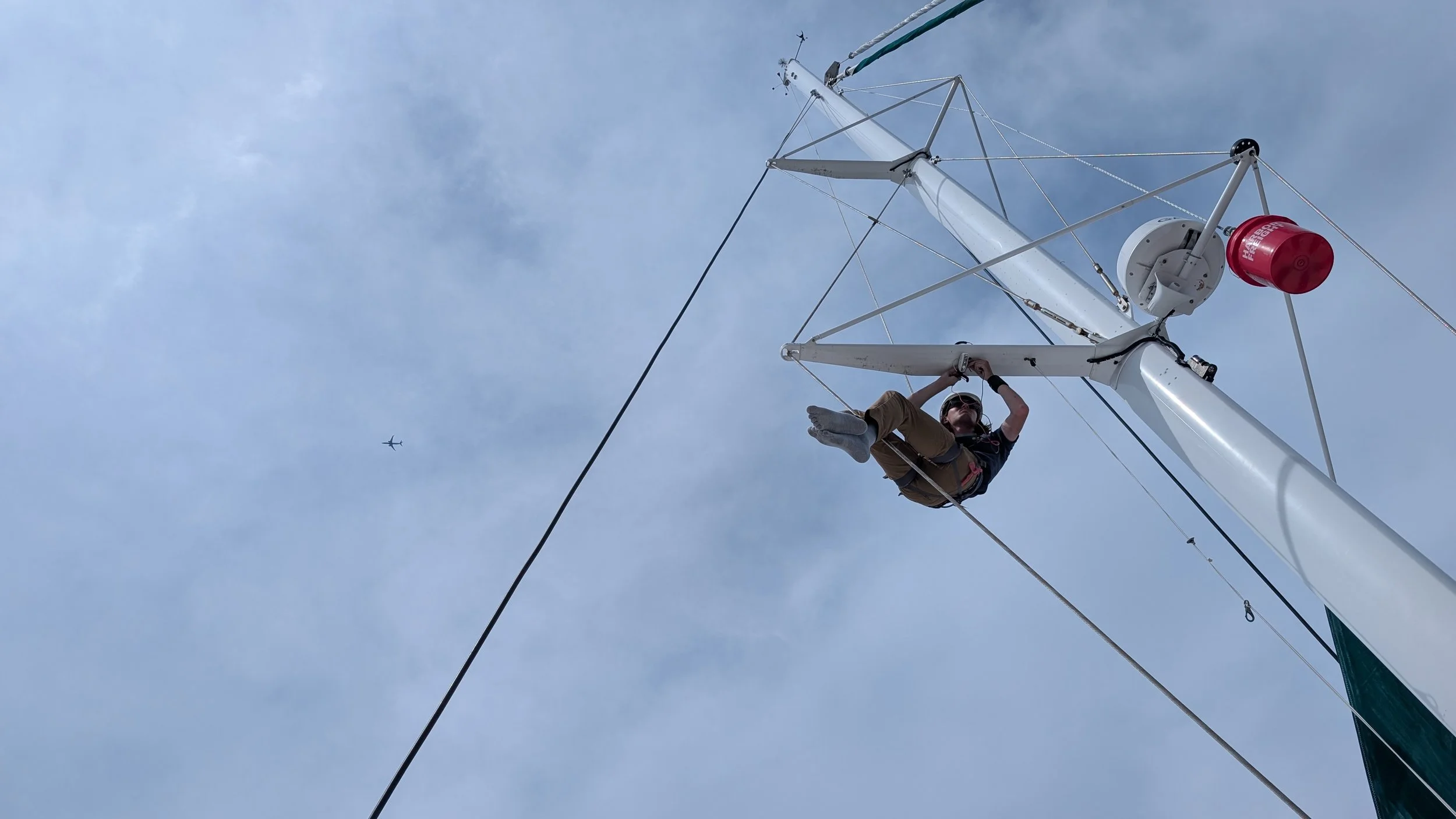 A person hanging upside down on a sailboat mast with gray sky background and a small airplane flying in the distance.