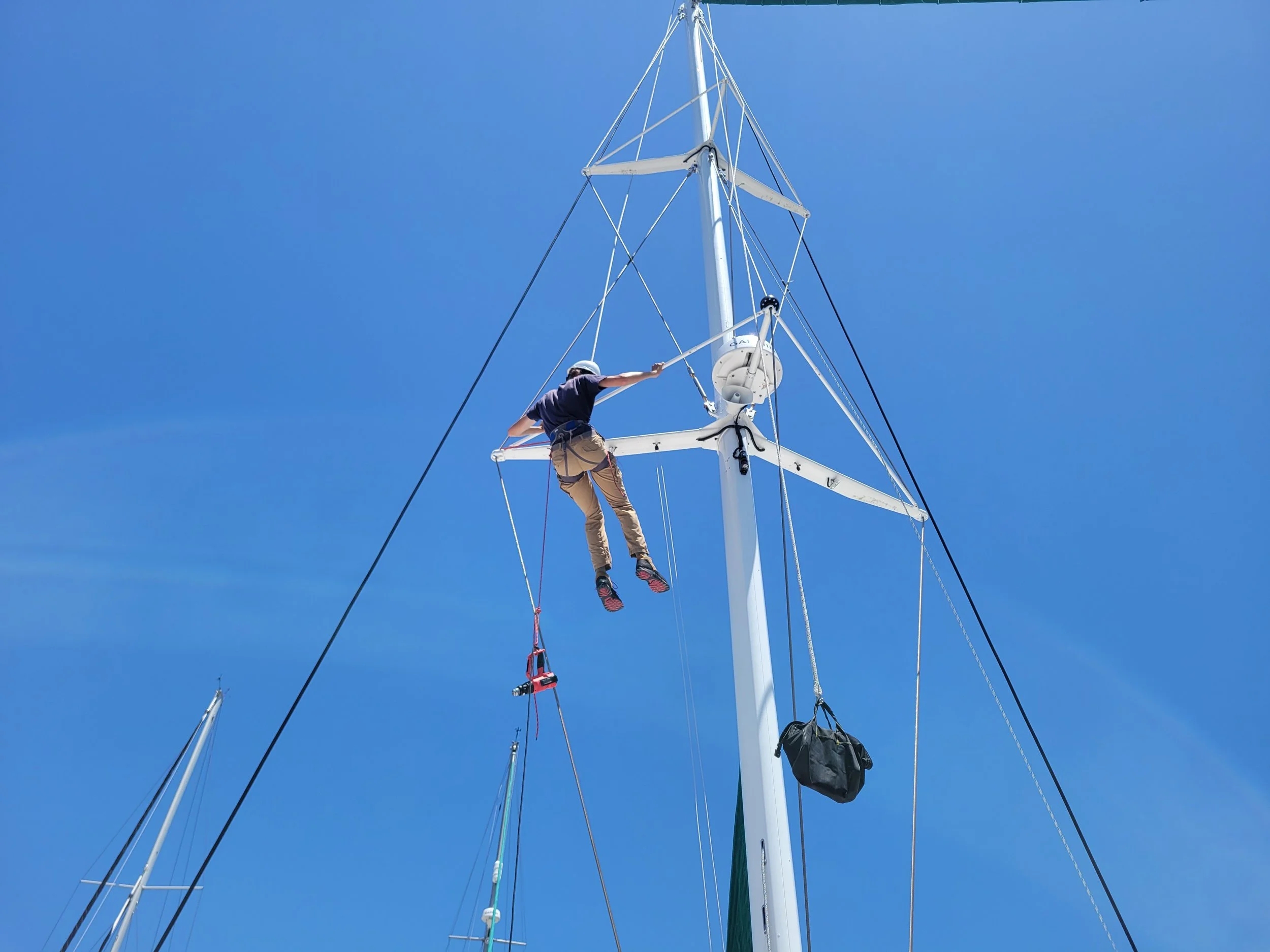 A person wearing a helmet and safety harness is cleaning the mast of a sailboat against a clear blue sky.