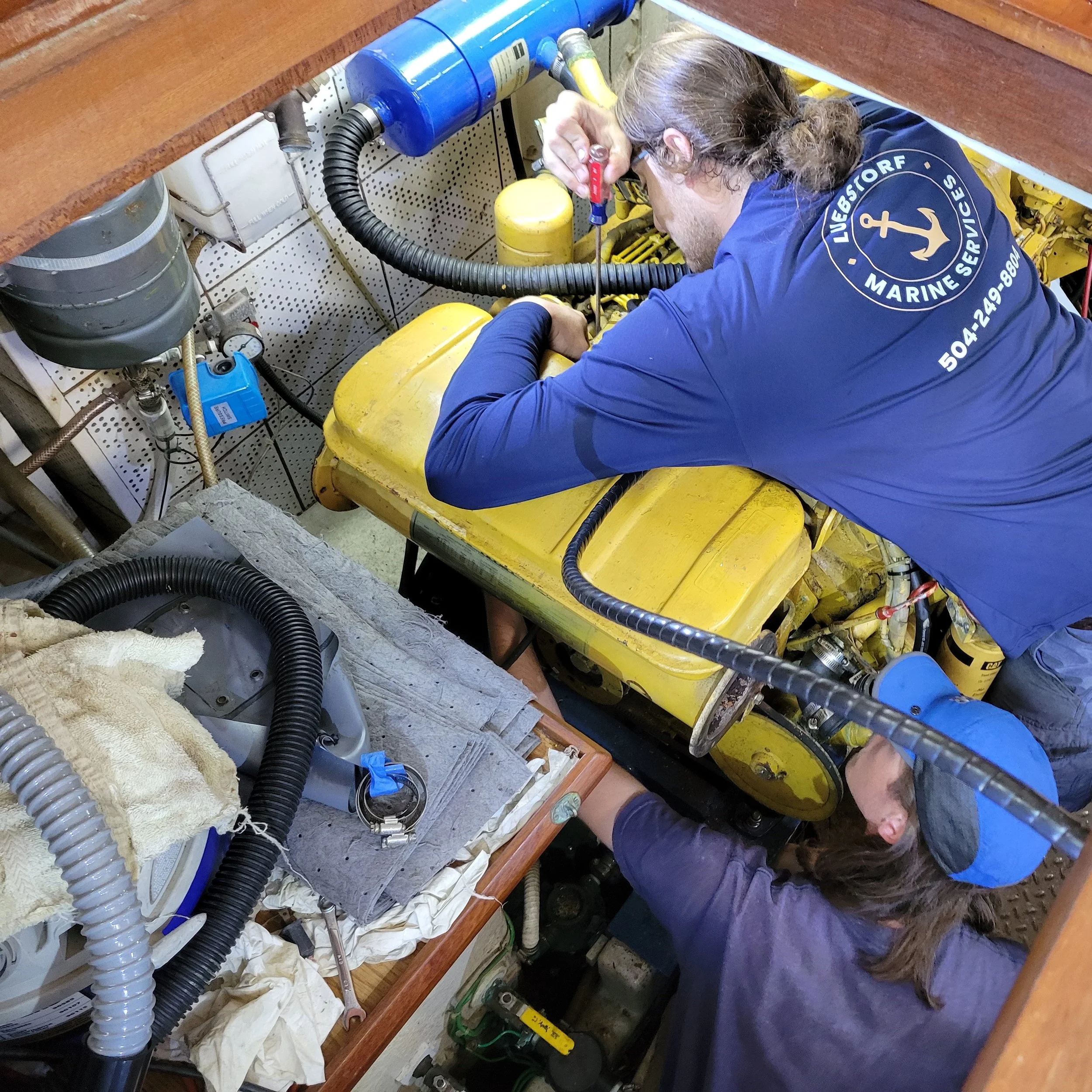 Two people working on a yellow marine engine inside a boat, with tools and equipment around.