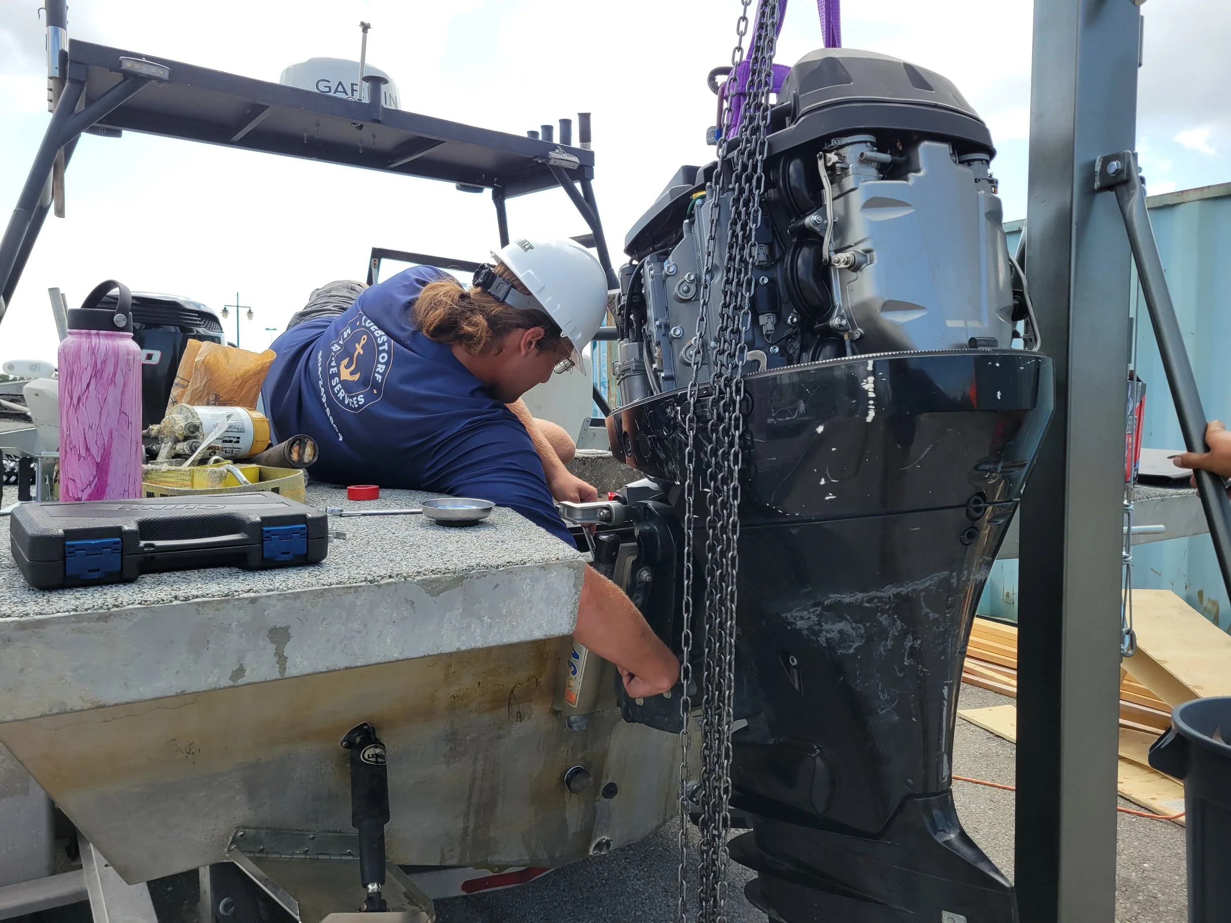 A person wearing a blue shirt and white hard hat working on the engine of a boat, which is mounted on a trailer. The work area has tools, a pink water bottle, and other equipment nearby.