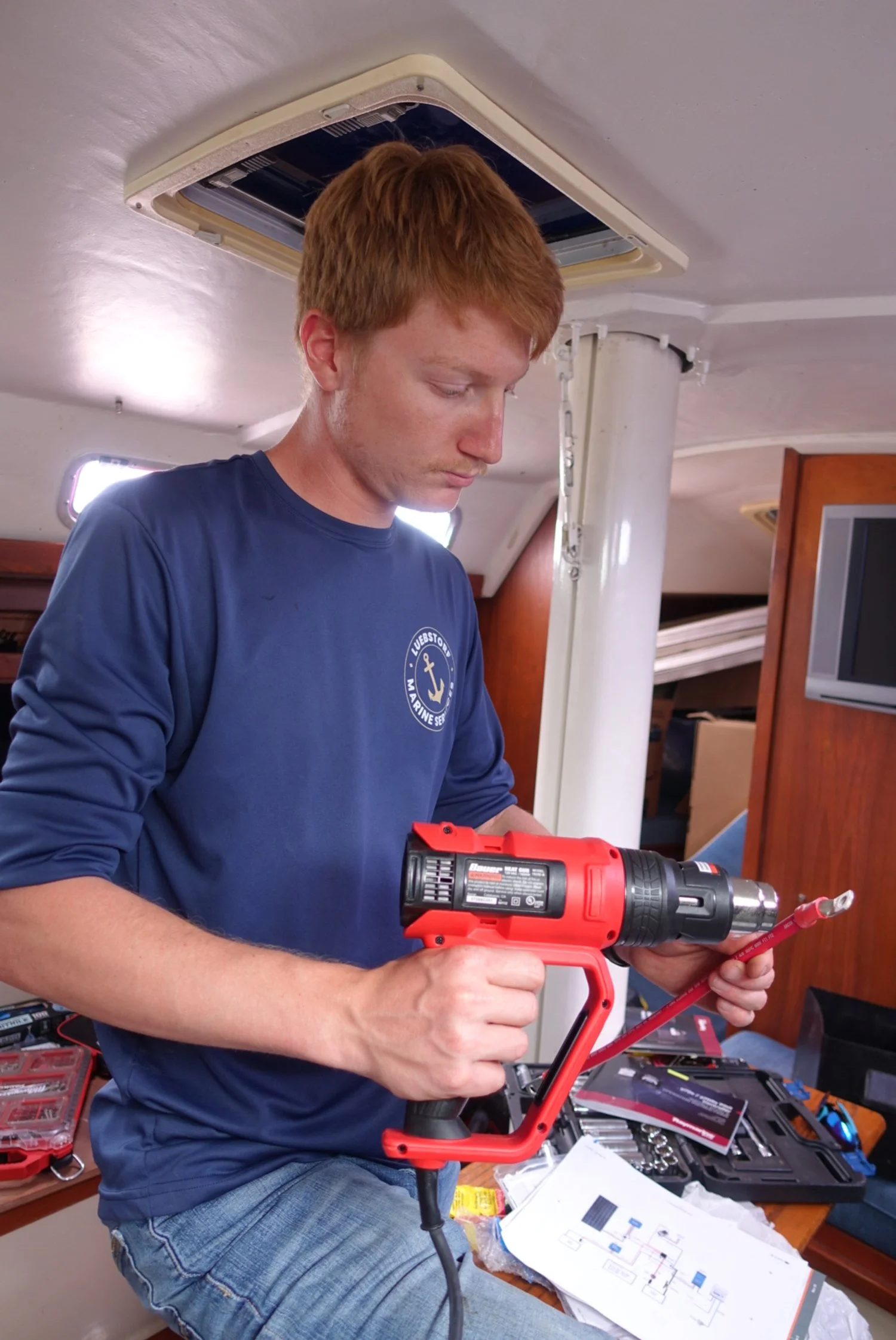 A man with red hair and a navy blue shirt using a red cordless drill on a boat's interior.