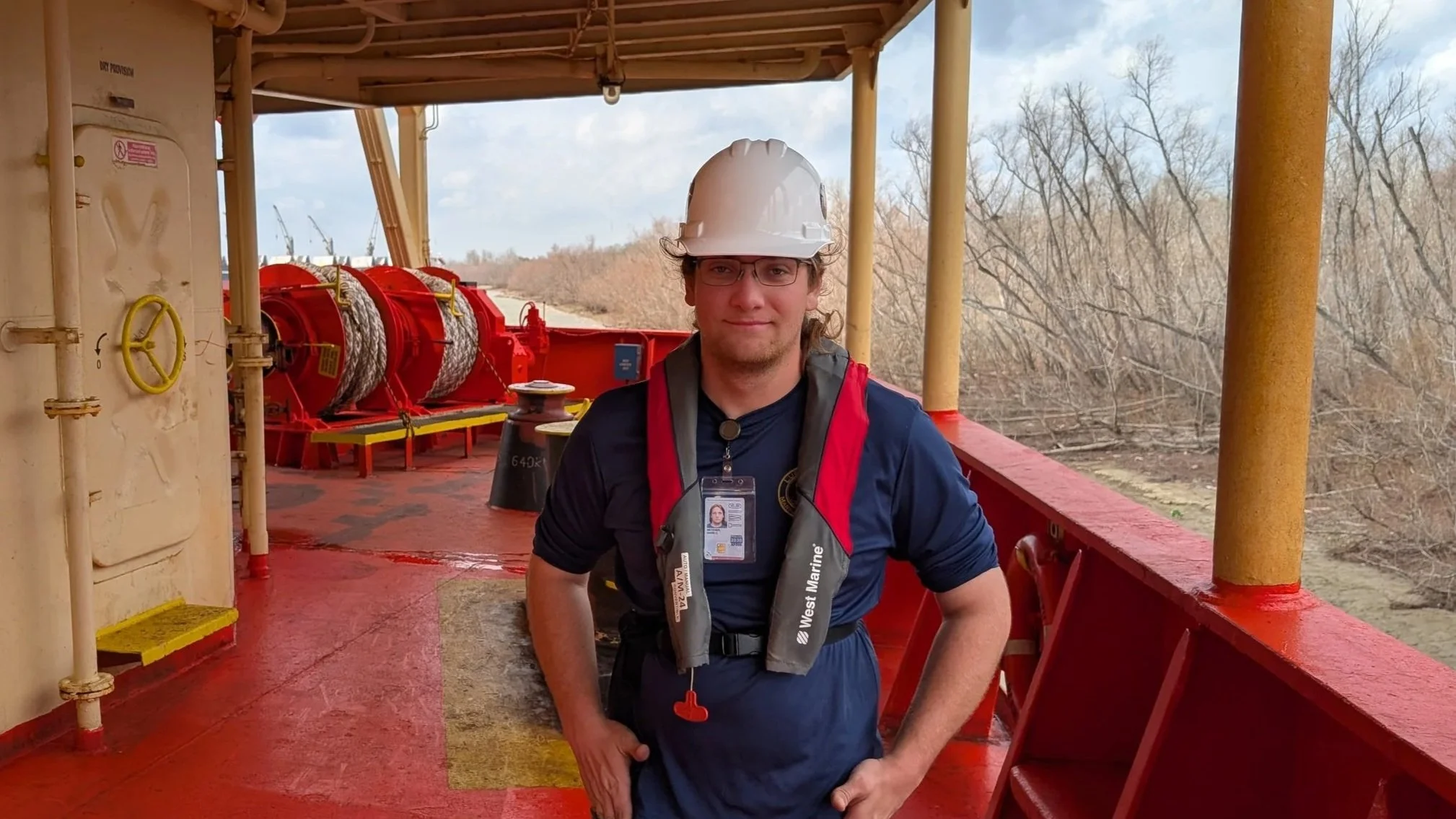 A person with long brown hair wearing glasses and a light green polo shirt, standing in front of a white door.