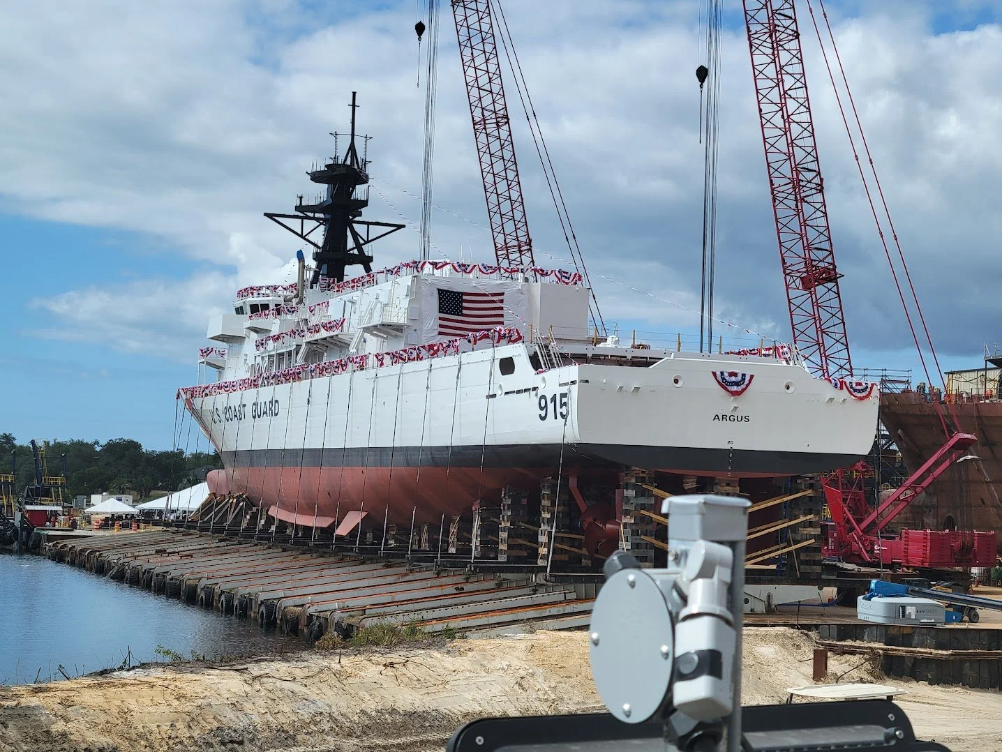 A large Coast Guard ship called 'Argus' on a dry dock for maintenance, decorated with patriotic flags and bunting, with cranes and construction equipment around.
