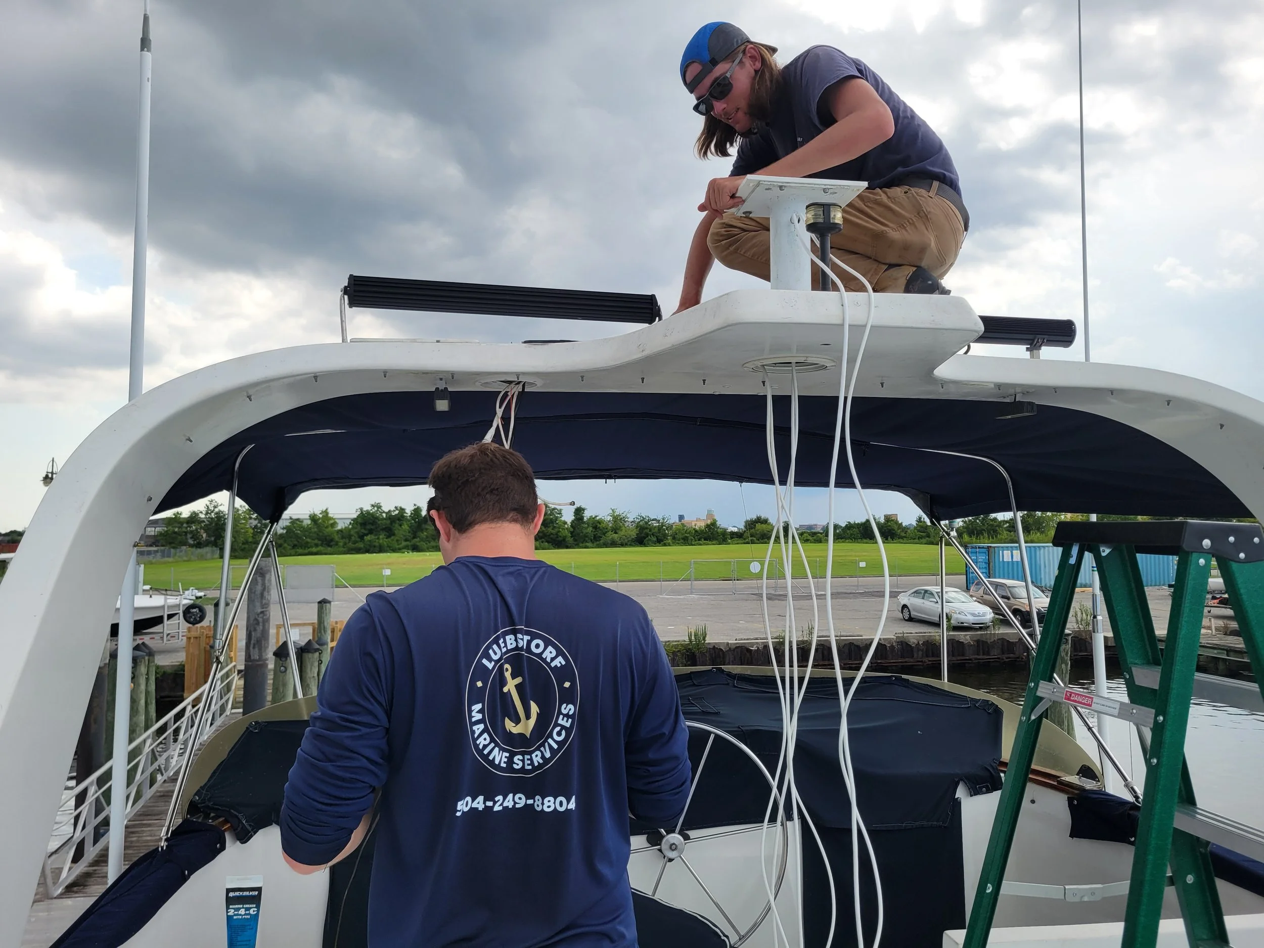 Two men working on a boat dock with one on top and one below, surrounded by cloudy sky and a green field in the background.