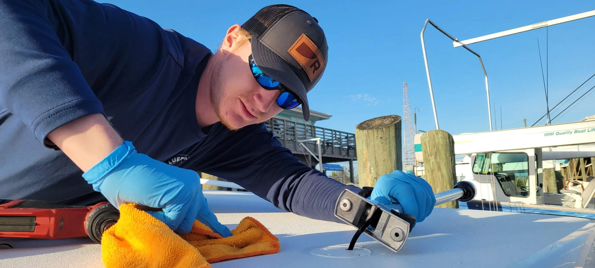 Man wearing a black cap, blue sunglasses, blue gloves, and a dark long sleeve shirt working on a boat's surface with a wrench, with a towel nearby, at a marina during daytime.
