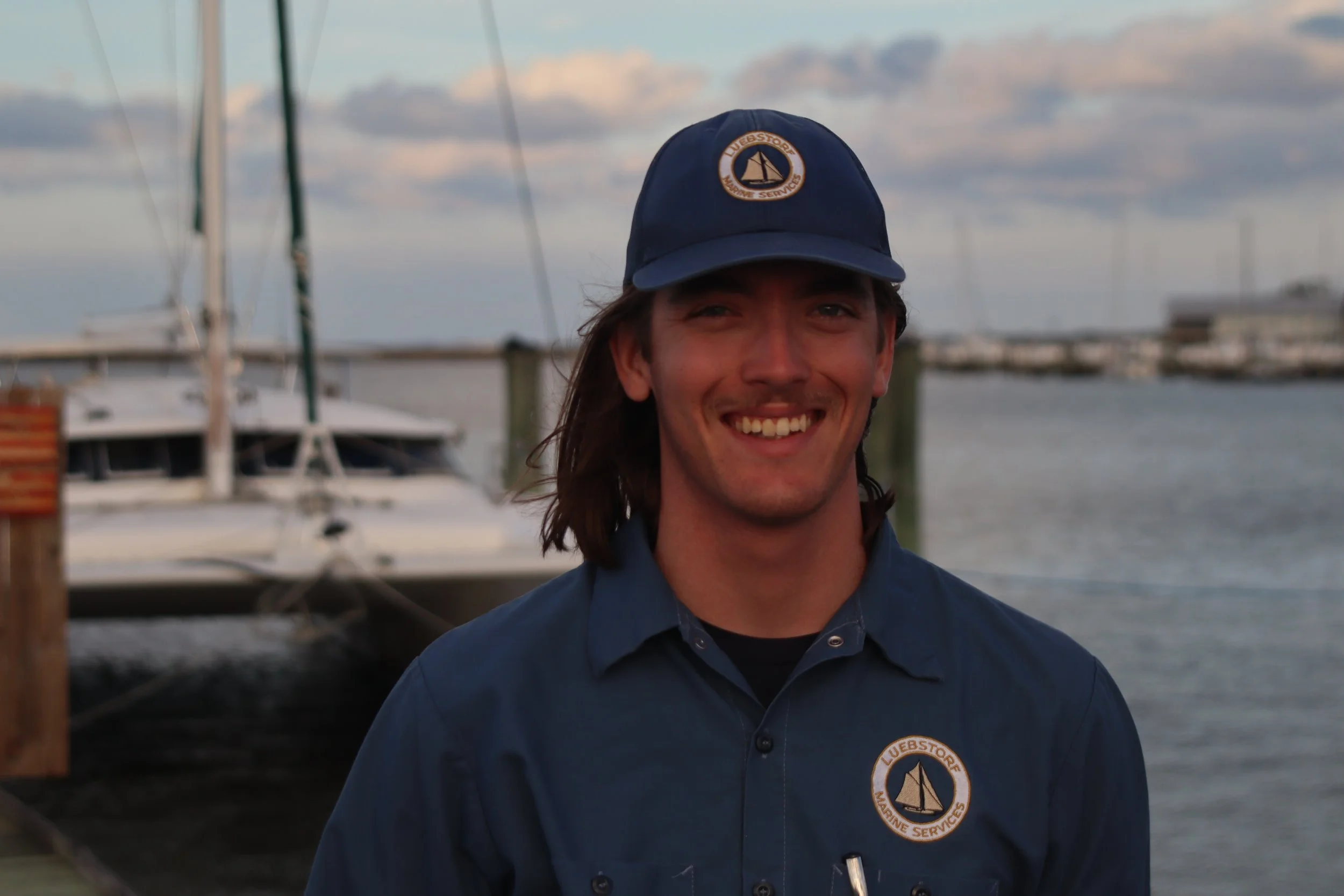A young man with long brown hair, glasses, and a light blue shirt smiling outdoors.