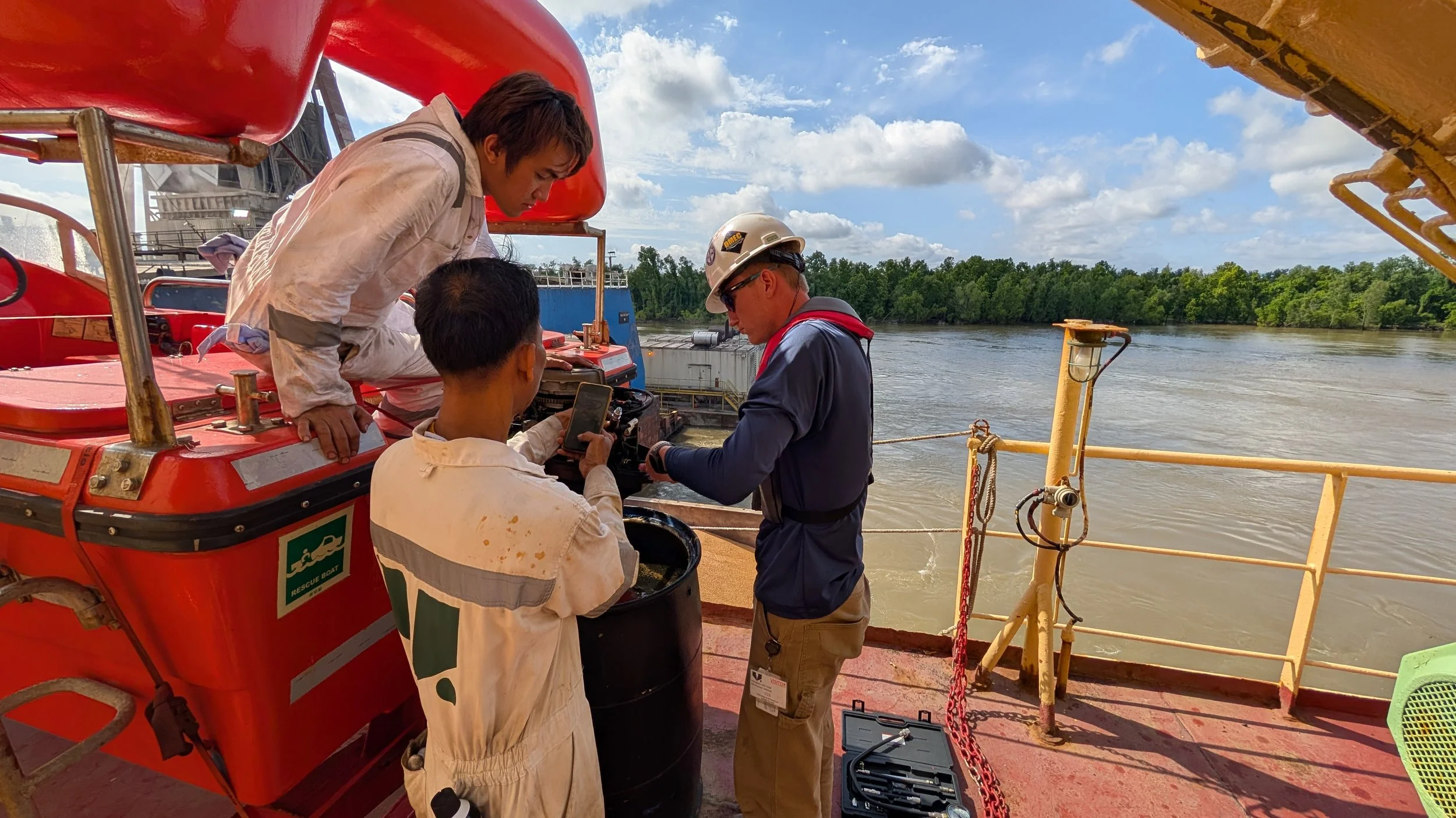 Three workers on a ship examining equipment near the water, with trees and a partly cloudy sky in the background.