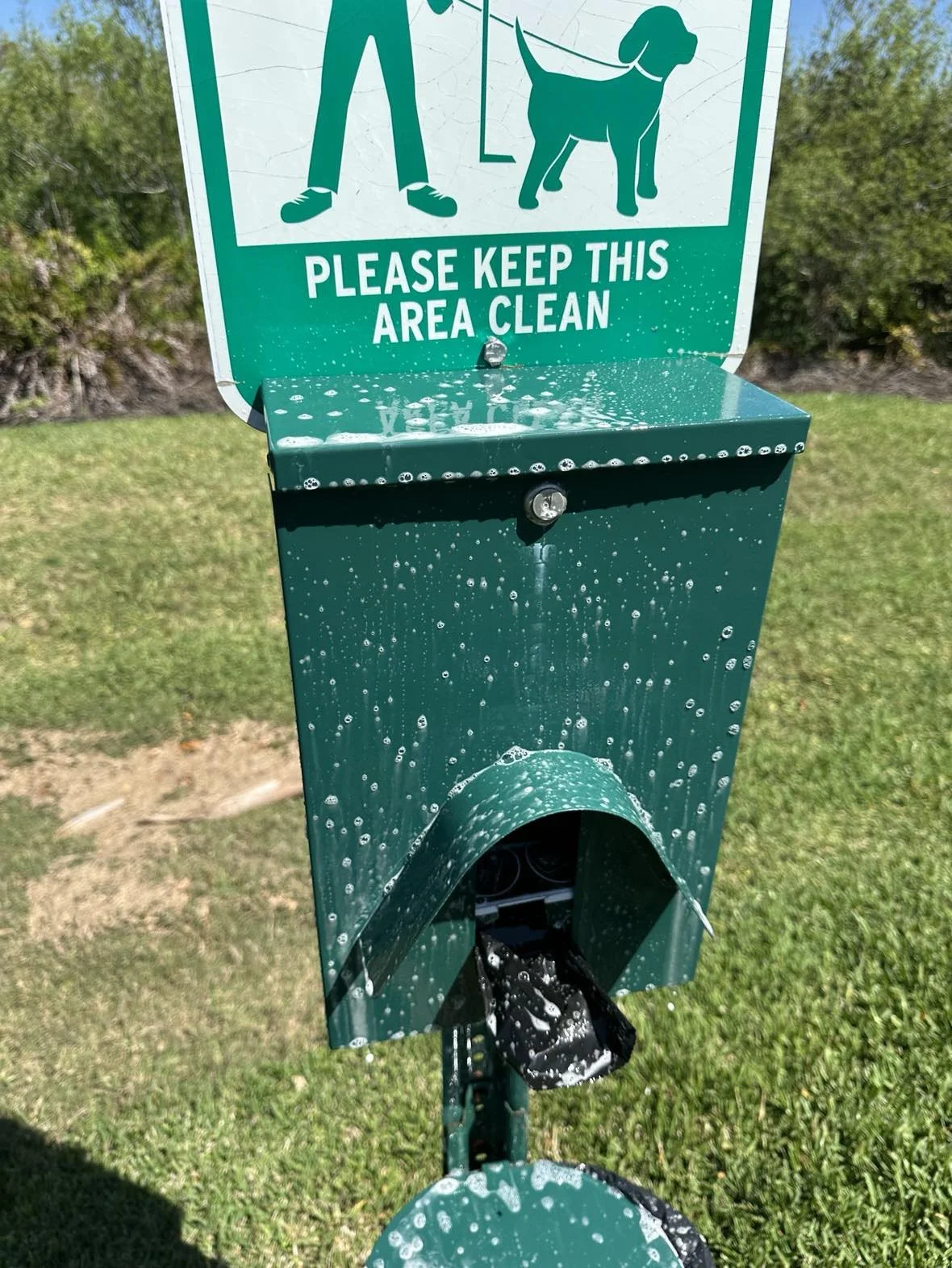 Dog waste station with a sign to keep the area clean, water on the station, and a waste bag dispenser. The station is green with a graphic of a person walking a dog.