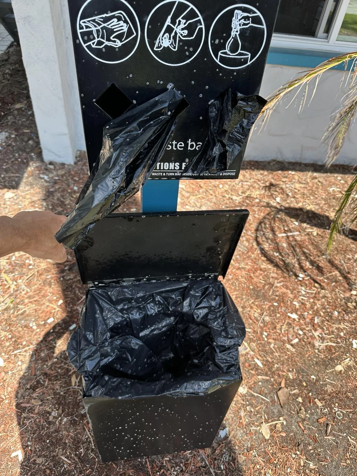 A person is holding a black trash bag over a black trash bin with a starry night design, located outdoors near a waste disposal station with instructional signs. The signs depict steps for waste disposal.