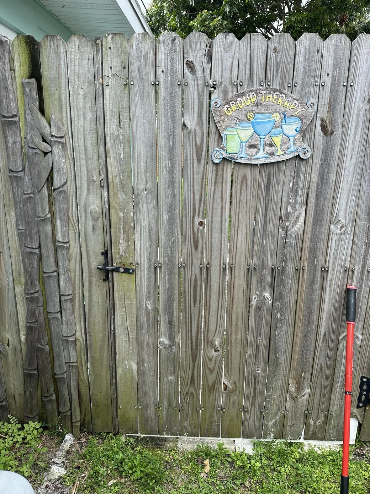 Wooden gate with a metal latch, decorated with a sign that reads 'Group Therapy' with images of colorful cocktails, and a garden tool leaning against the right side.