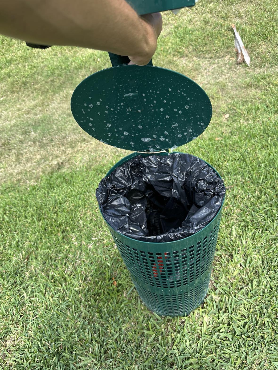A green outdoor trash can with a black plastic liner inside, open on a grassy area. The can's lid is propped open, and water droplets are visible on the inside of the lid.