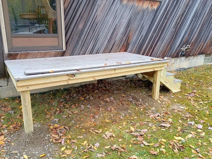 Wooden deck with stairs attached to the side of a house, situated on a grassy area with fallen leaves.