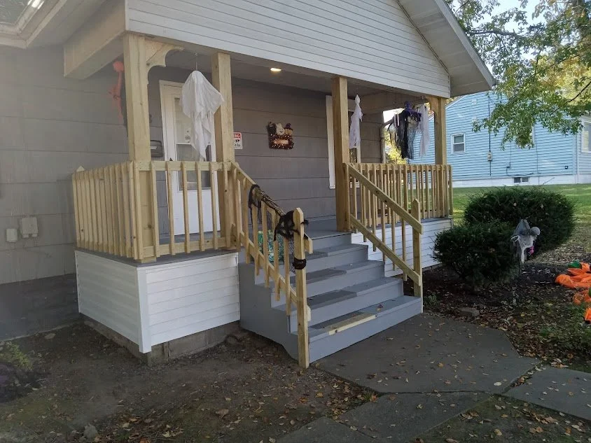 Front porch of a house with a newly built wooden staircase and railing, Halloween decorations including ghosts hanging on the porch, and a small garden with bushes and some Halloween-themed decorations on the ground.
