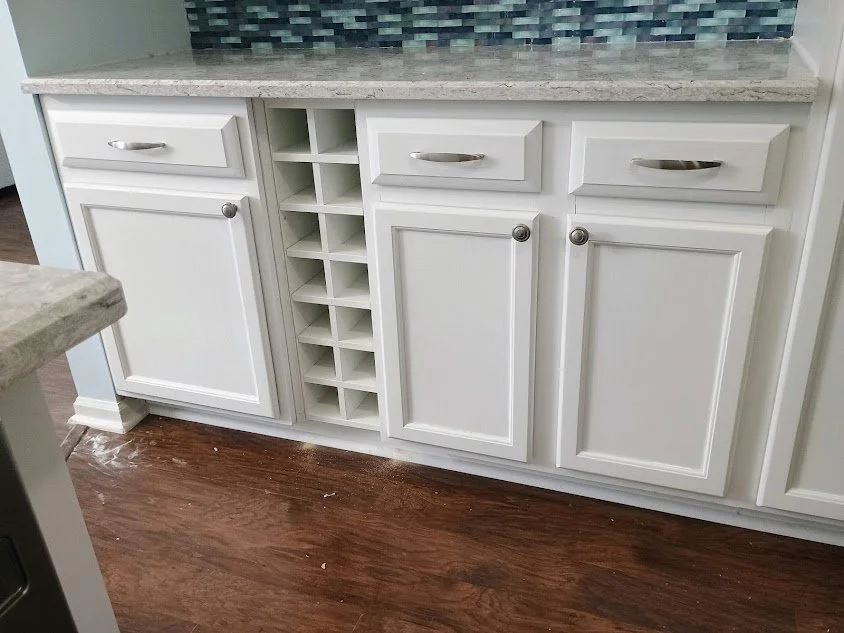 White kitchen cabinet with multiple drawers, a small wine rack, and a granite countertop above, set against a tiled backsplash and hardwood floor.