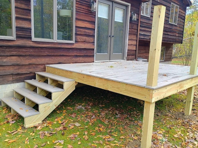 Wooden porch under construction with stairs leading to the door, attached to a brown house with windows, surrounded by fallen leaves.