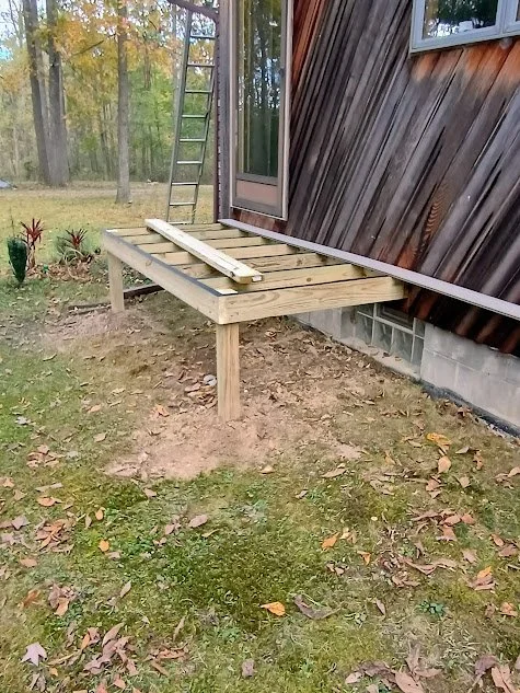 A wooden deck under construction next to a house, with a ladder leaning against the house and a few plants nearby.