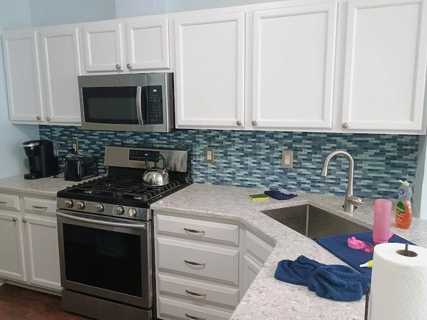 Kitchen with white cabinets, a stainless steel stove with a kettle on top, a microwave above the stove, blue tile backsplash, a black coffee maker, a sink with a pink tumbler and dish soap, blue dish drying mat, paper towel roll, and some cleaning cl