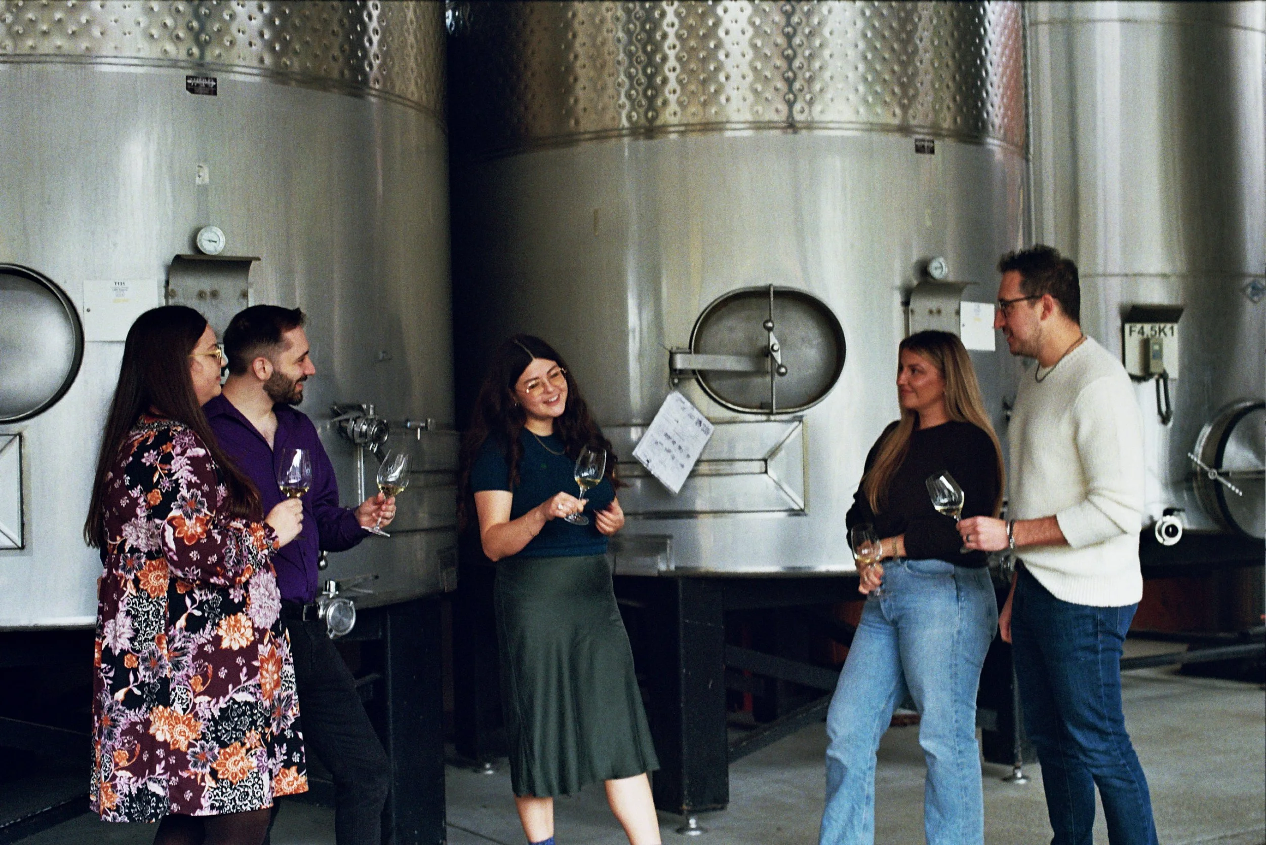 A tour group stands in front of fermentation tanks listening to their wine tour guide.