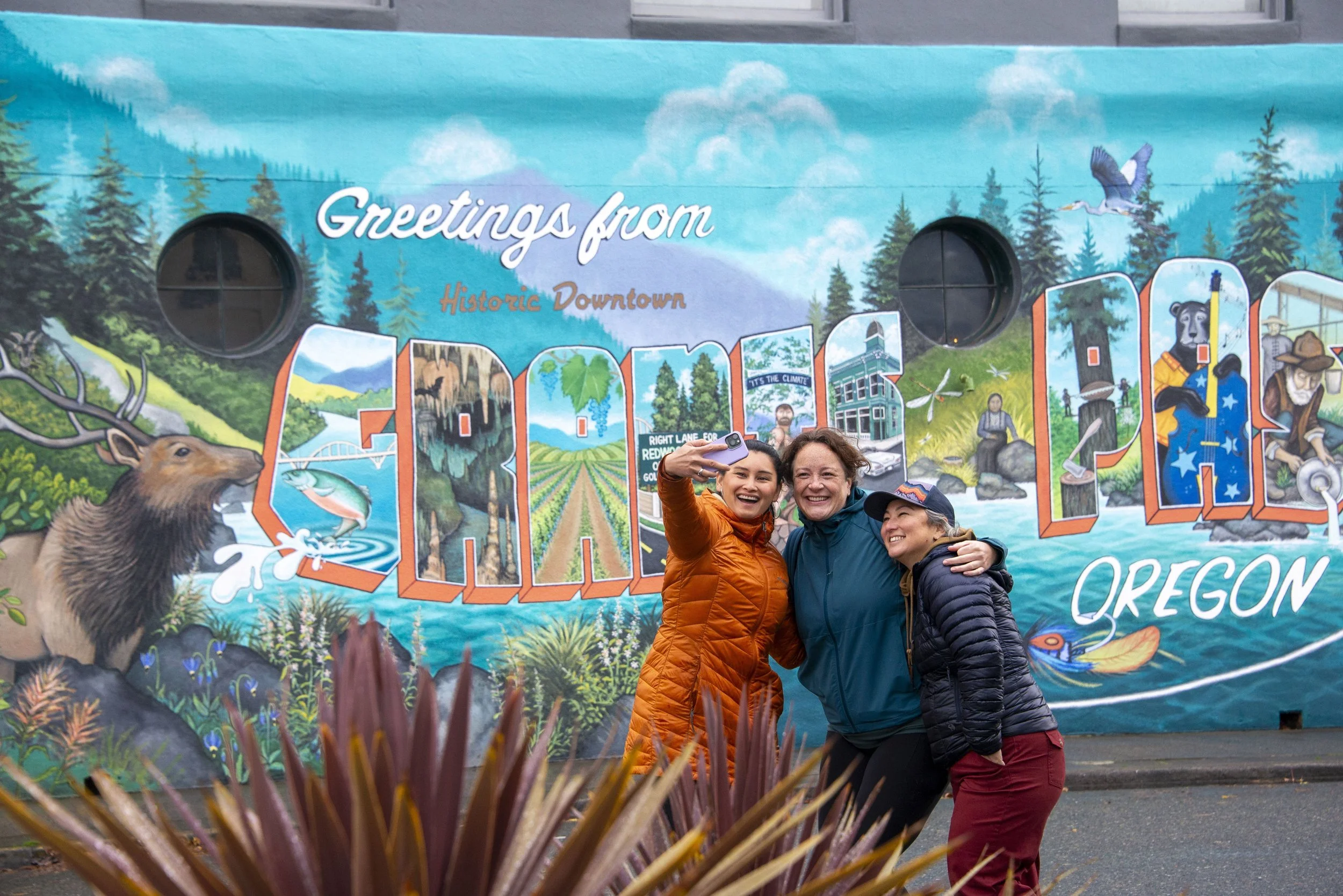 A group of three people take a selfie in front of a mural that says "Greetings from Grants Pass", one of the many things to do in Grants Pass.