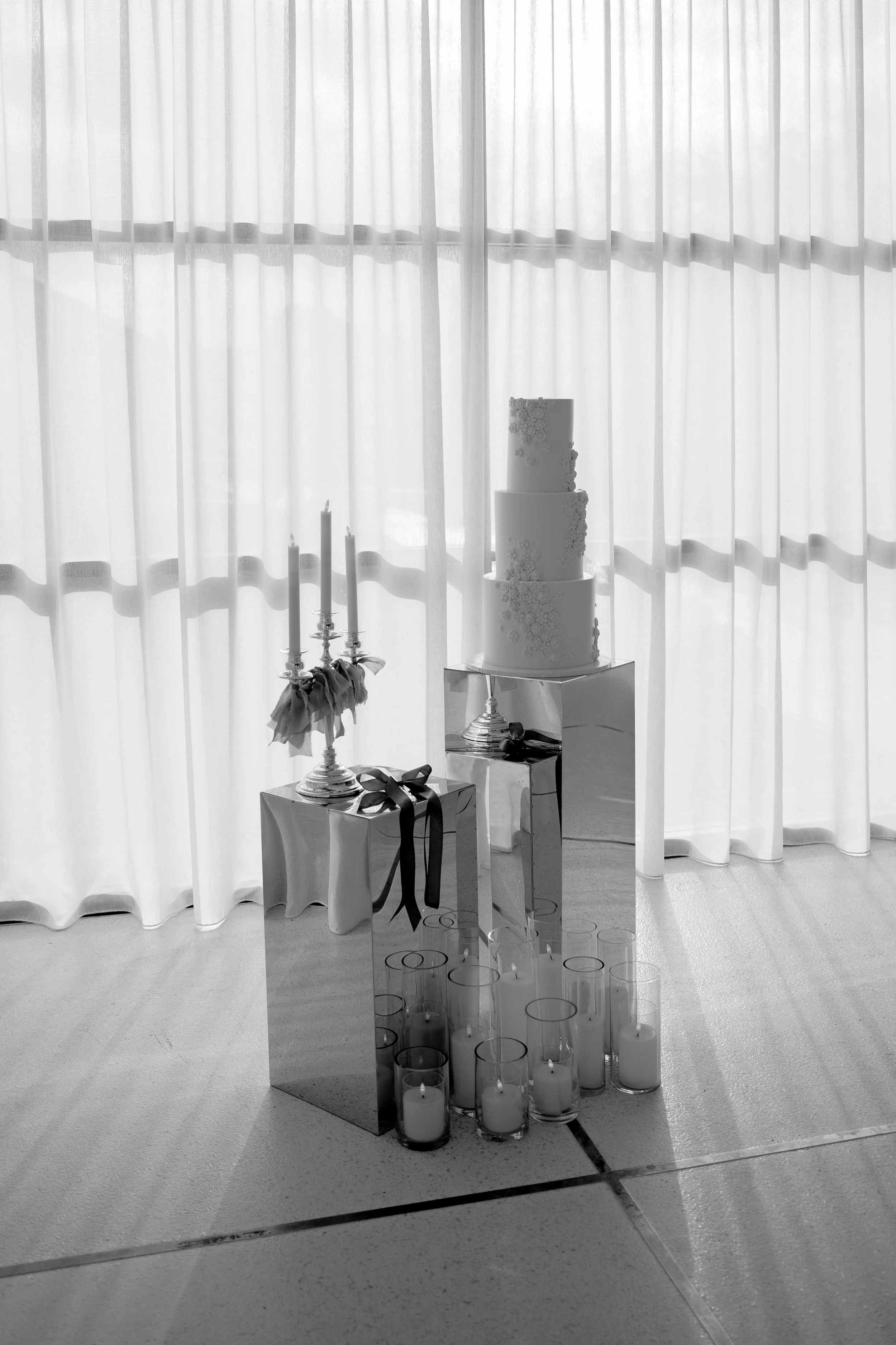 A wedding setup with a three-tier cake, candle holders with candles on mirrored tables, and glass candle holders arranged on the floor in front of a sheer curtain.