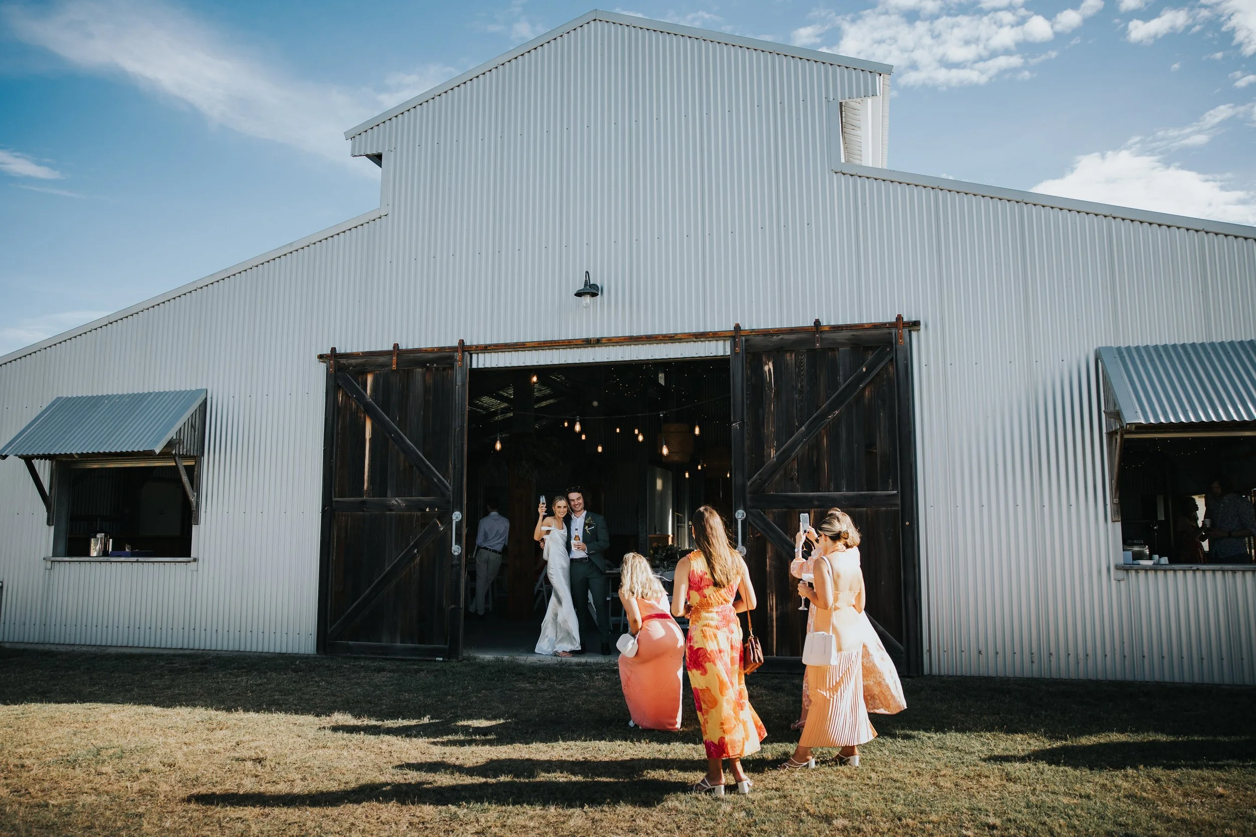 Guests celebrating at a wedding reception outside a rustic barn with sliding doors, under a partly cloudy sky.