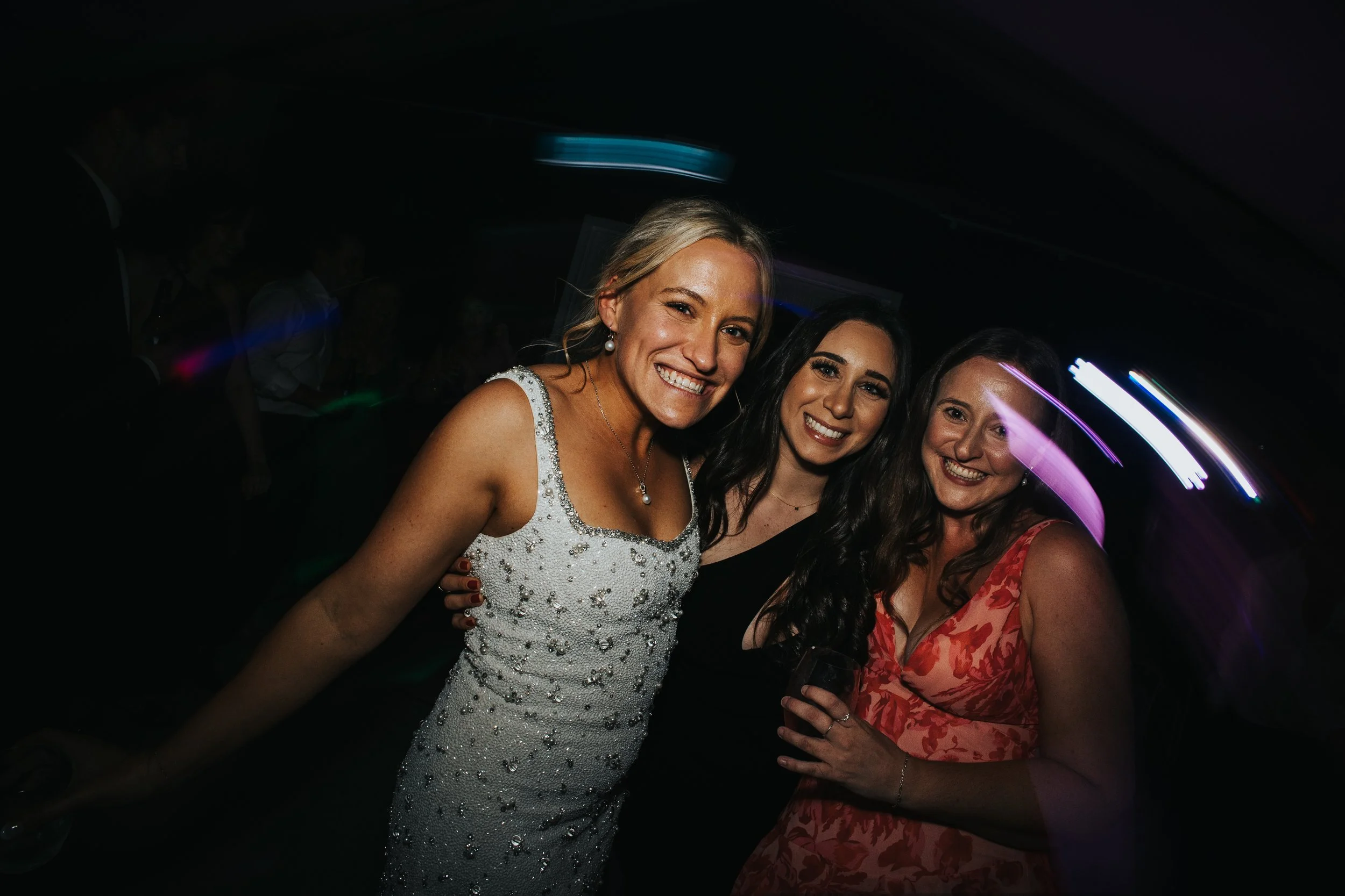 Group of three women smiling at a party with colorful lights in the background.