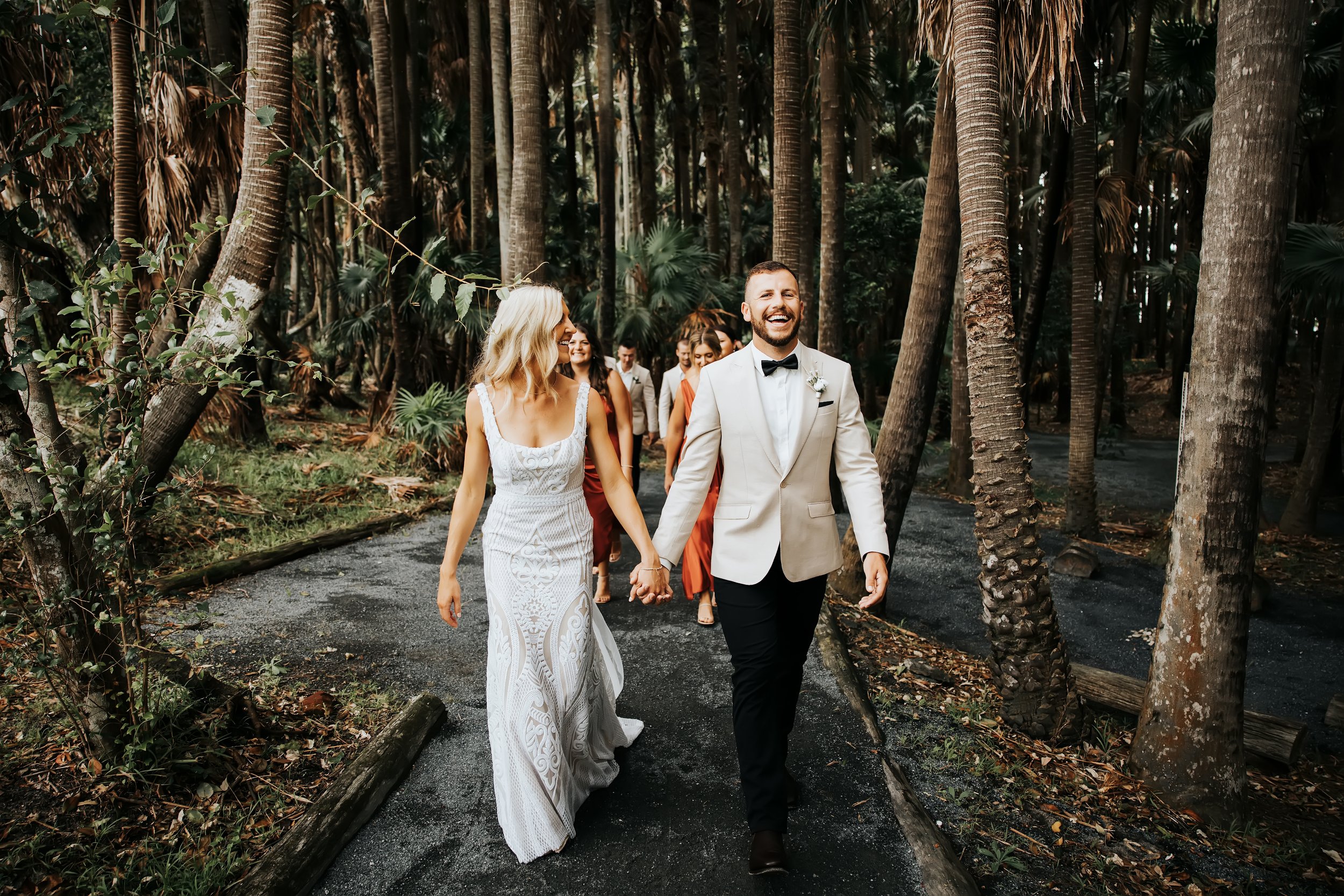 A wedding couple, a woman in a white lace wedding dress and a man in a white jacket with a black bow tie, holding hands and walking through a forest with tall trees and tropical plants, smiling and surrounded by friends or family.