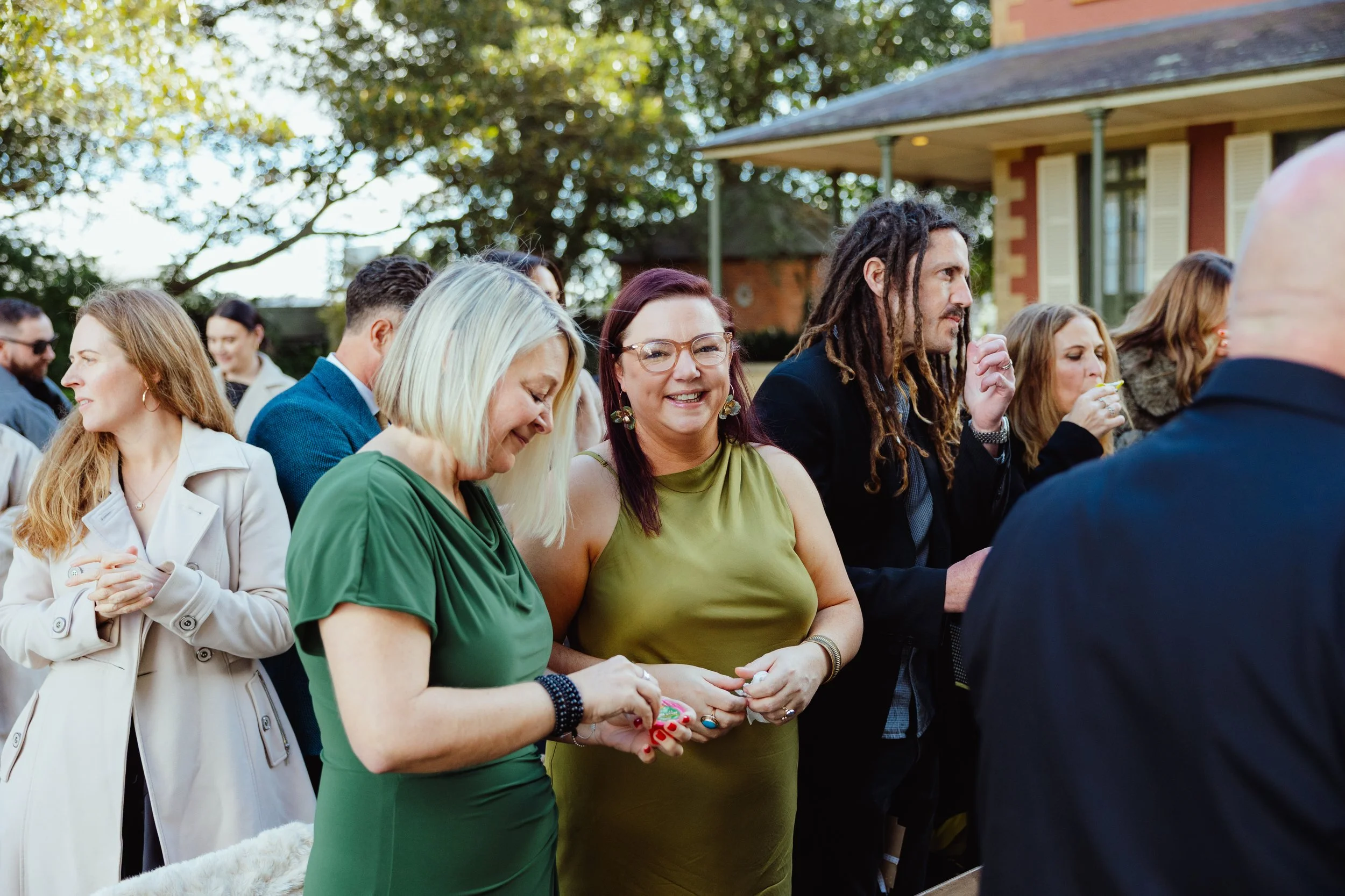 A group of people standing outdoors, enjoying a social gathering in front of a house with trees in the background.