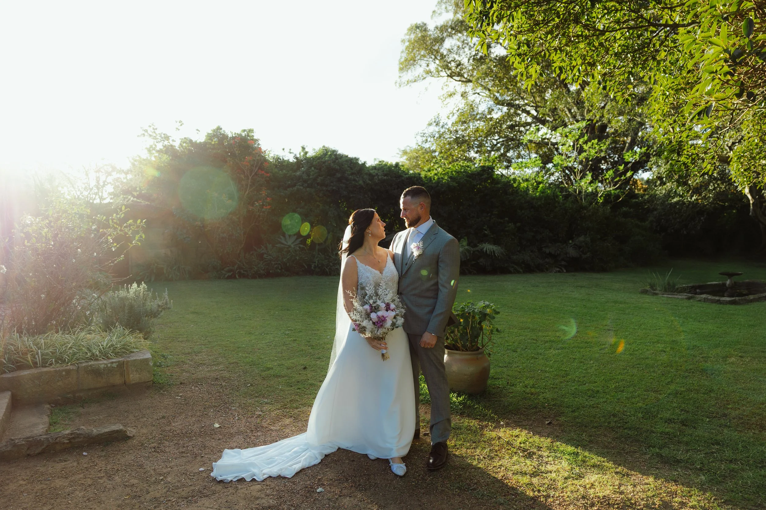 A bride and groom stand together in a lush garden during their wedding, with sunlight filtering through the trees.