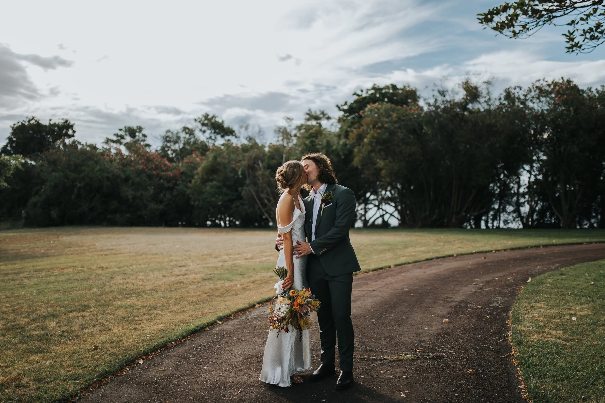 A bride and groom share a kiss outdoors on a cloudy day, surrounded by trees and open grass.