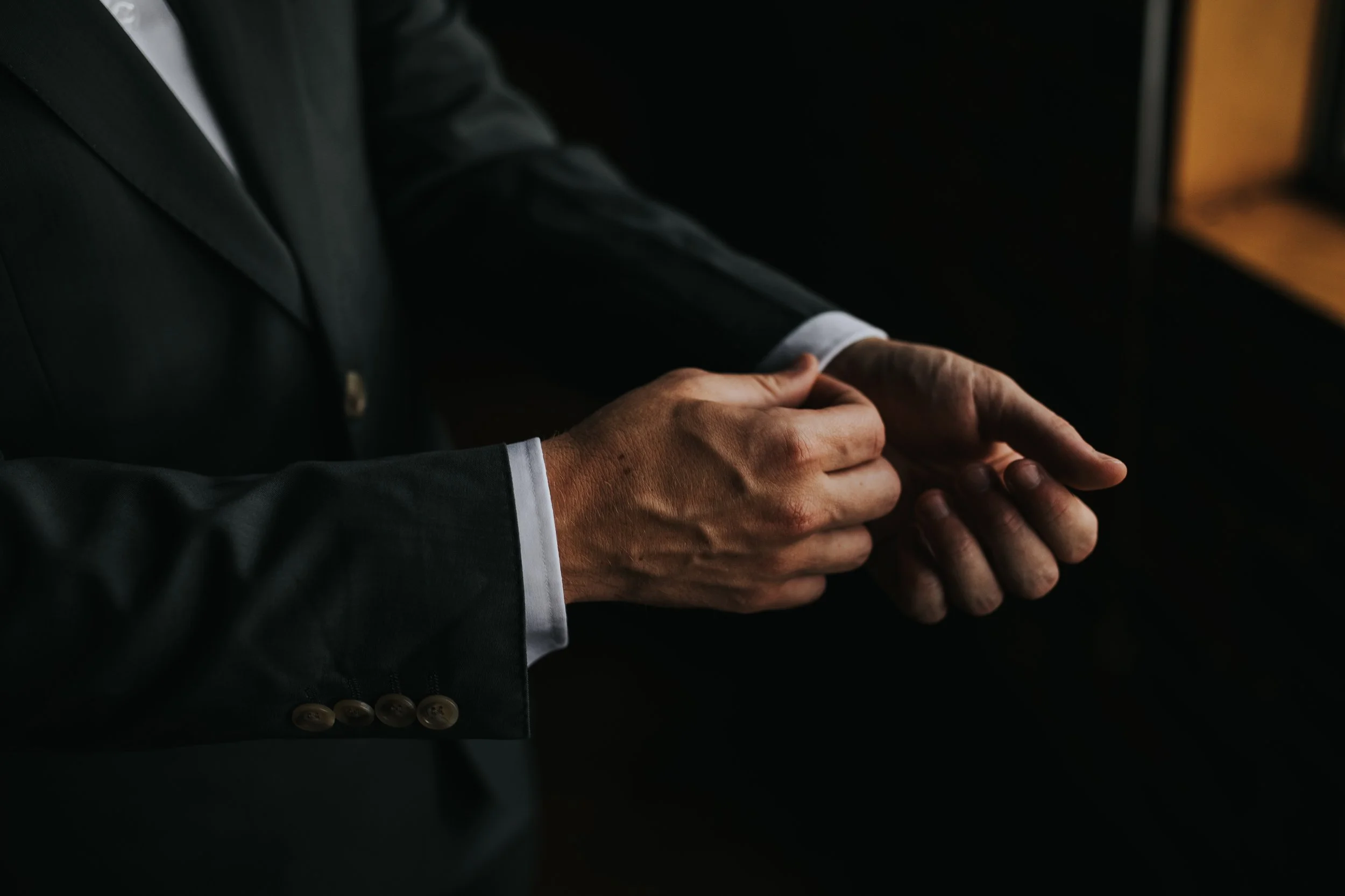 Close-up of two hands clasped together, wearing a dark suit with black buttons.