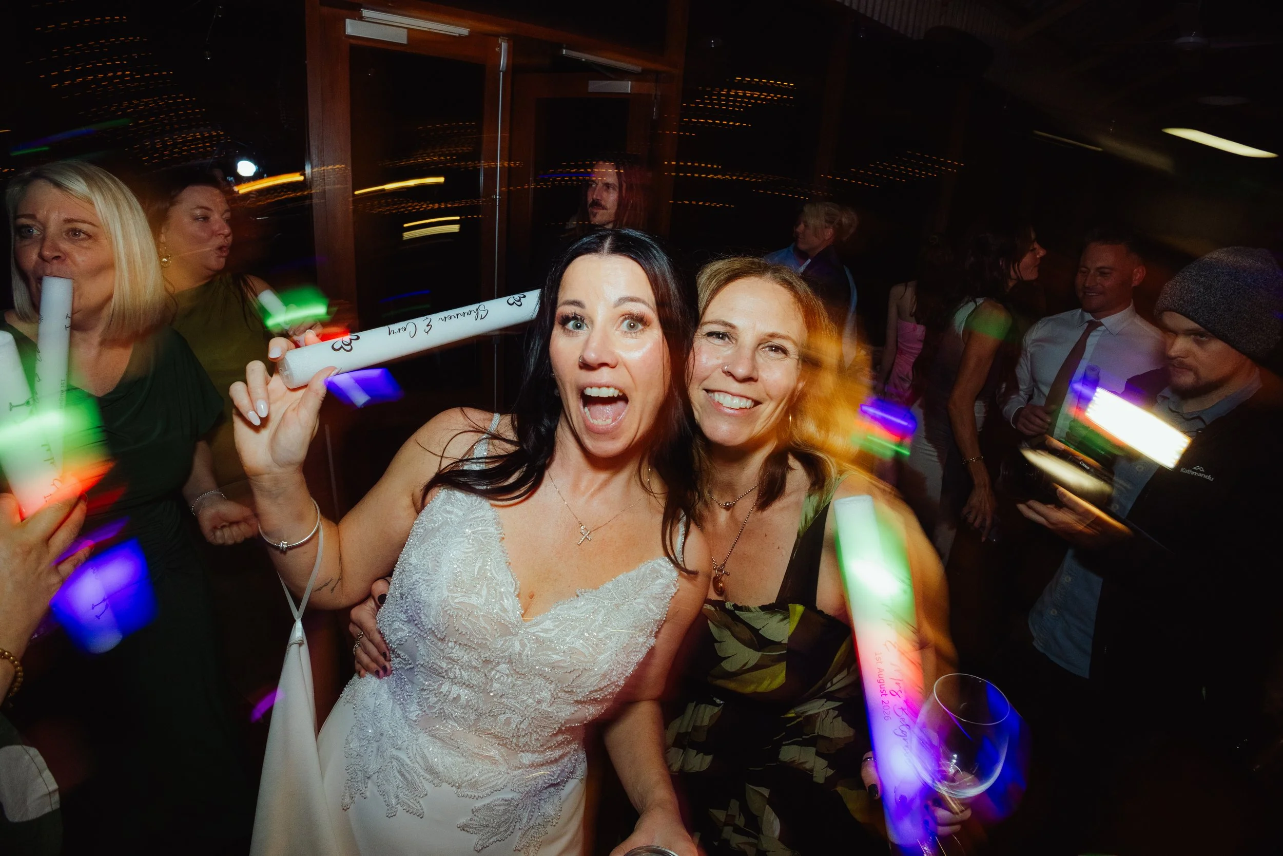 Group of people celebrating at a party, with two women in the foreground smiling and holding glow sticks, one wearing a white dress and the other in a floral dress, with other guests in the background under colorful lights.