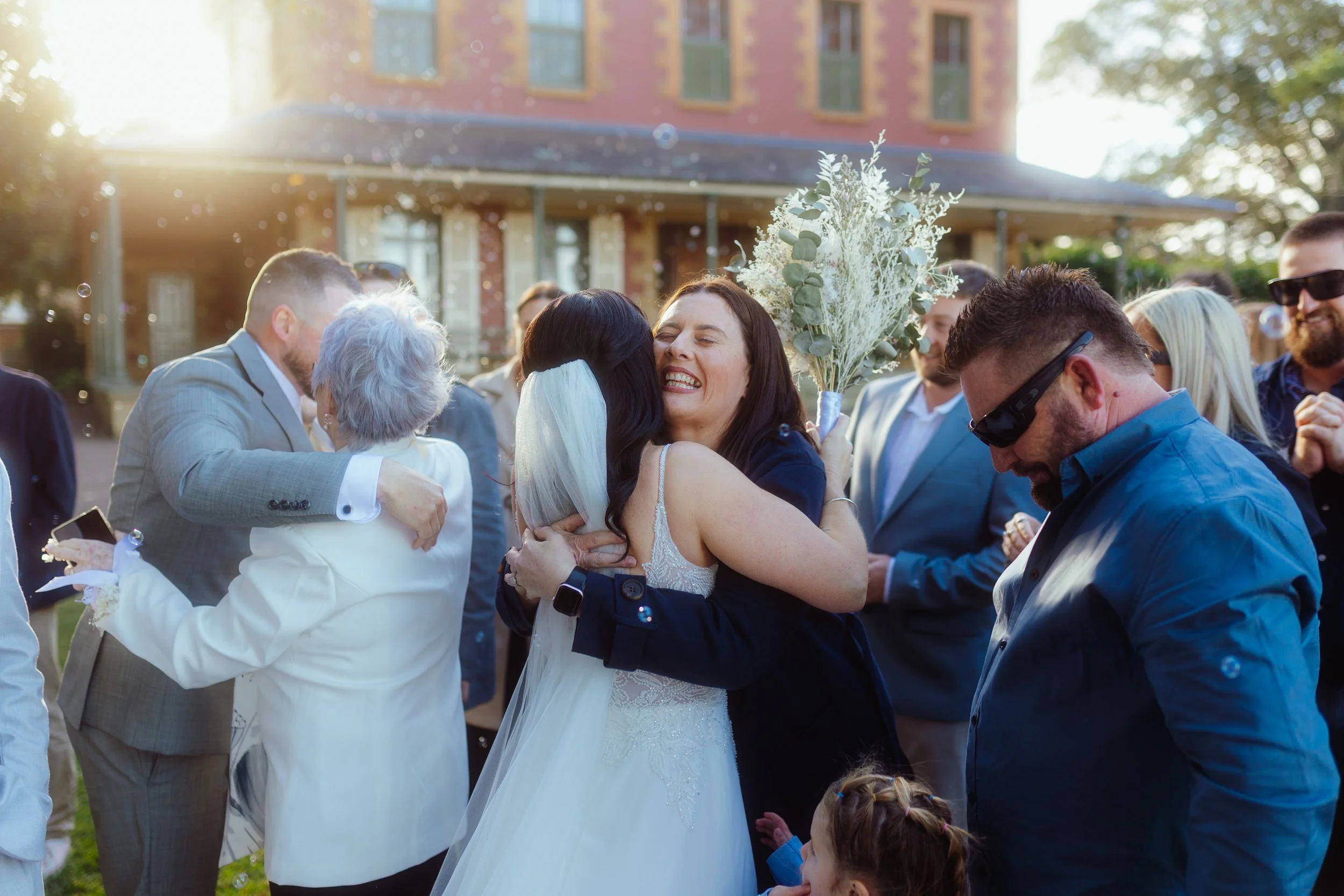 Group of people celebrating outdoors with hugs and smiles, including a woman in a wedding dress holding a bouquet, in front of a brick building with trees and sunlight in the background.