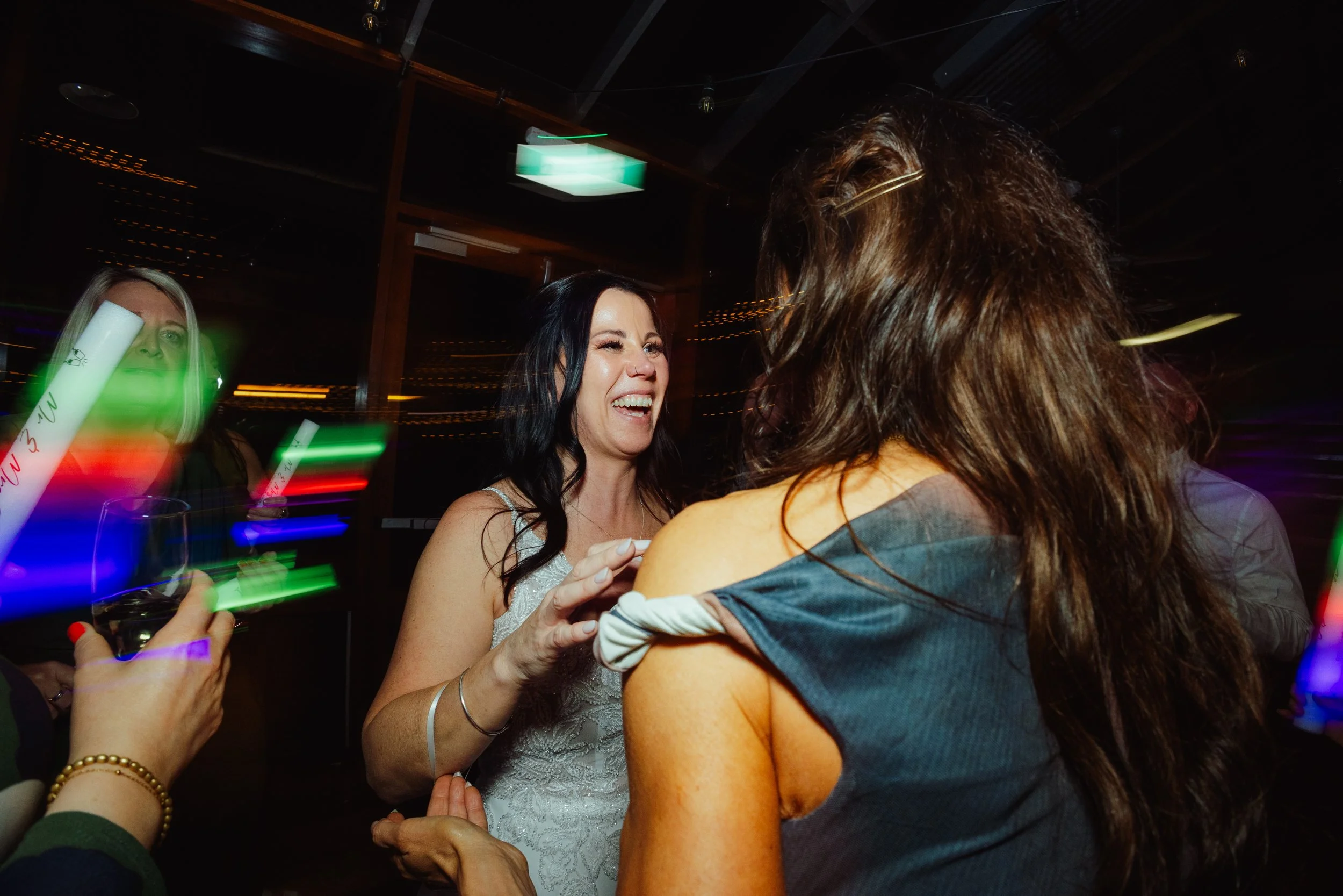 People at a lively party or celebration, smiling and dancing in a dimly lit room, with colorful lights and a fire exit sign visible.