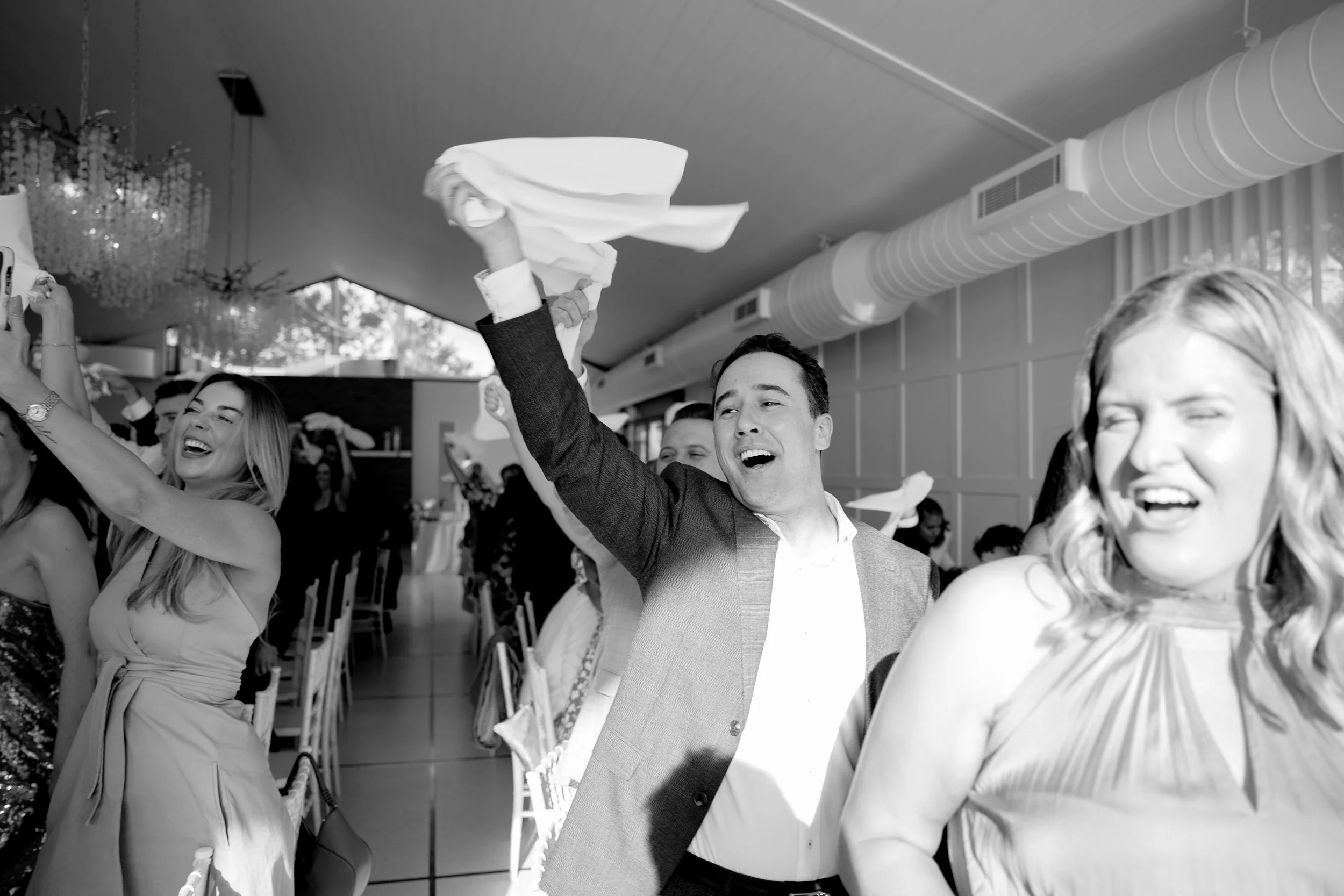 People celebrating at a wedding reception, cheering and raising their arms. A man in a suit is holding a cloth in the air, smiling, surrounded by women smiling and dancing.