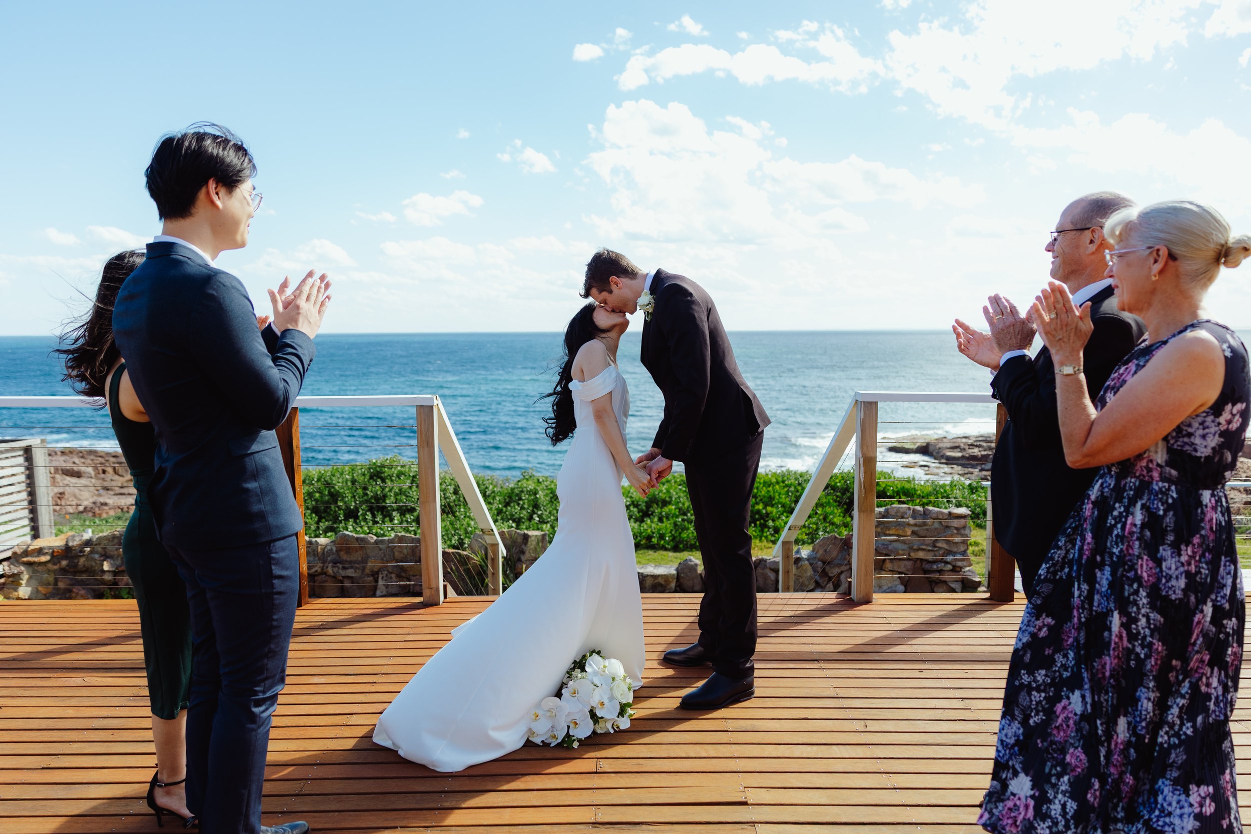 A couple gets married on a wooden deck overlooking the ocean with a clear sky, while guests stand and applaud.