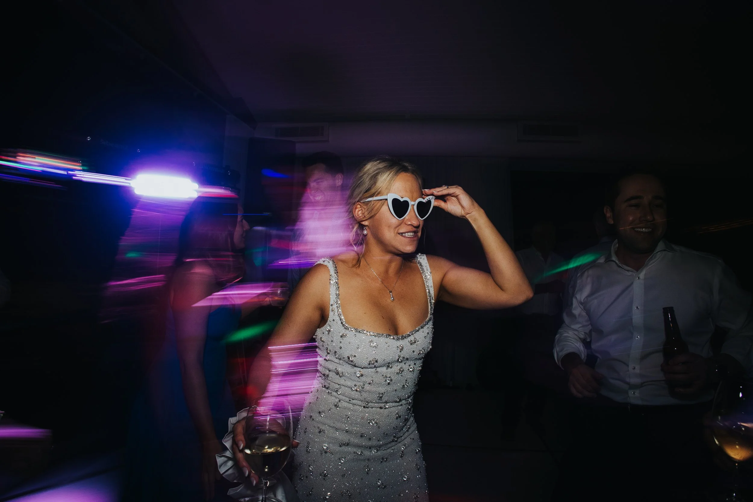 A woman wearing heart-shaped sunglasses and a sparkly dress dancing at a party, with colorful lights and other people in the background.