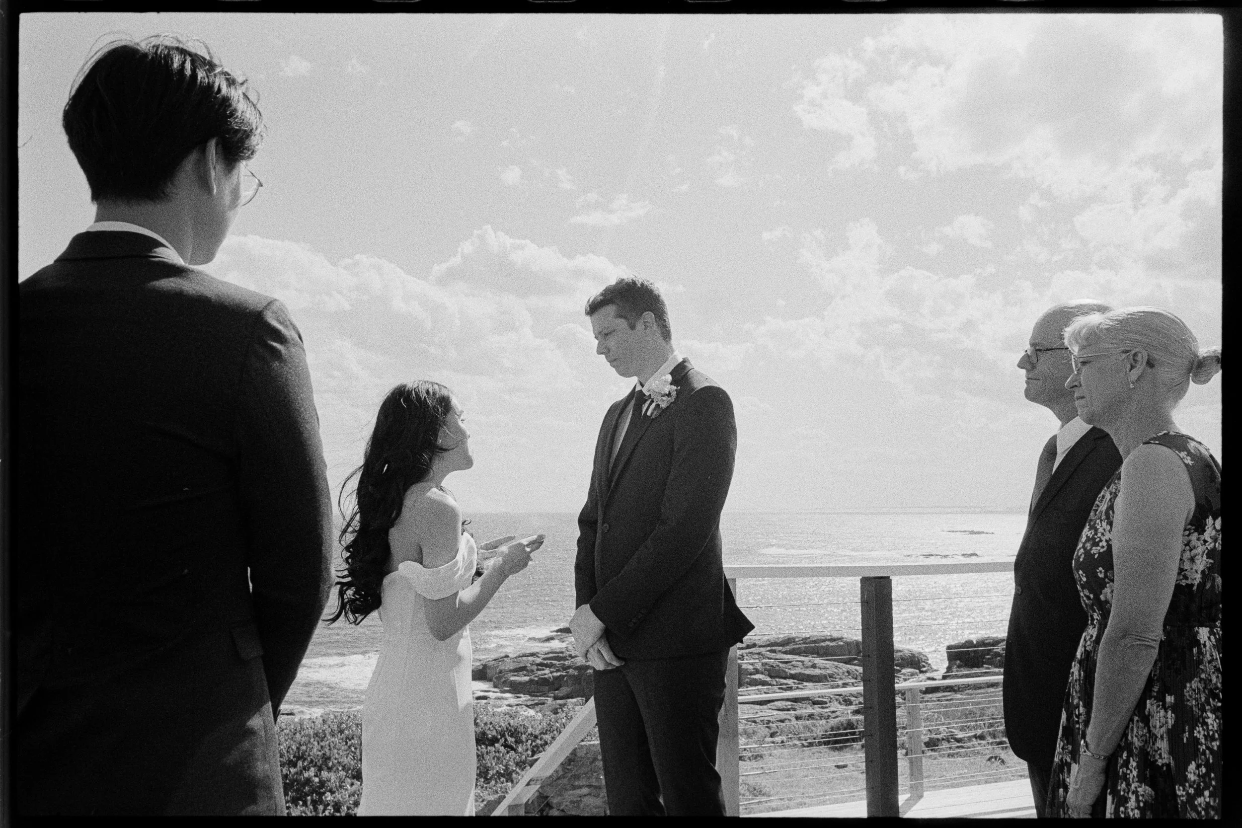 A wedding ceremony taking place on a beach, with six people standing in a row. The bride and groom are at the center, holding hands and exchanging vows. The bride is wearing a white dress, and the groom is wearing a suit. Two women and two men are st
