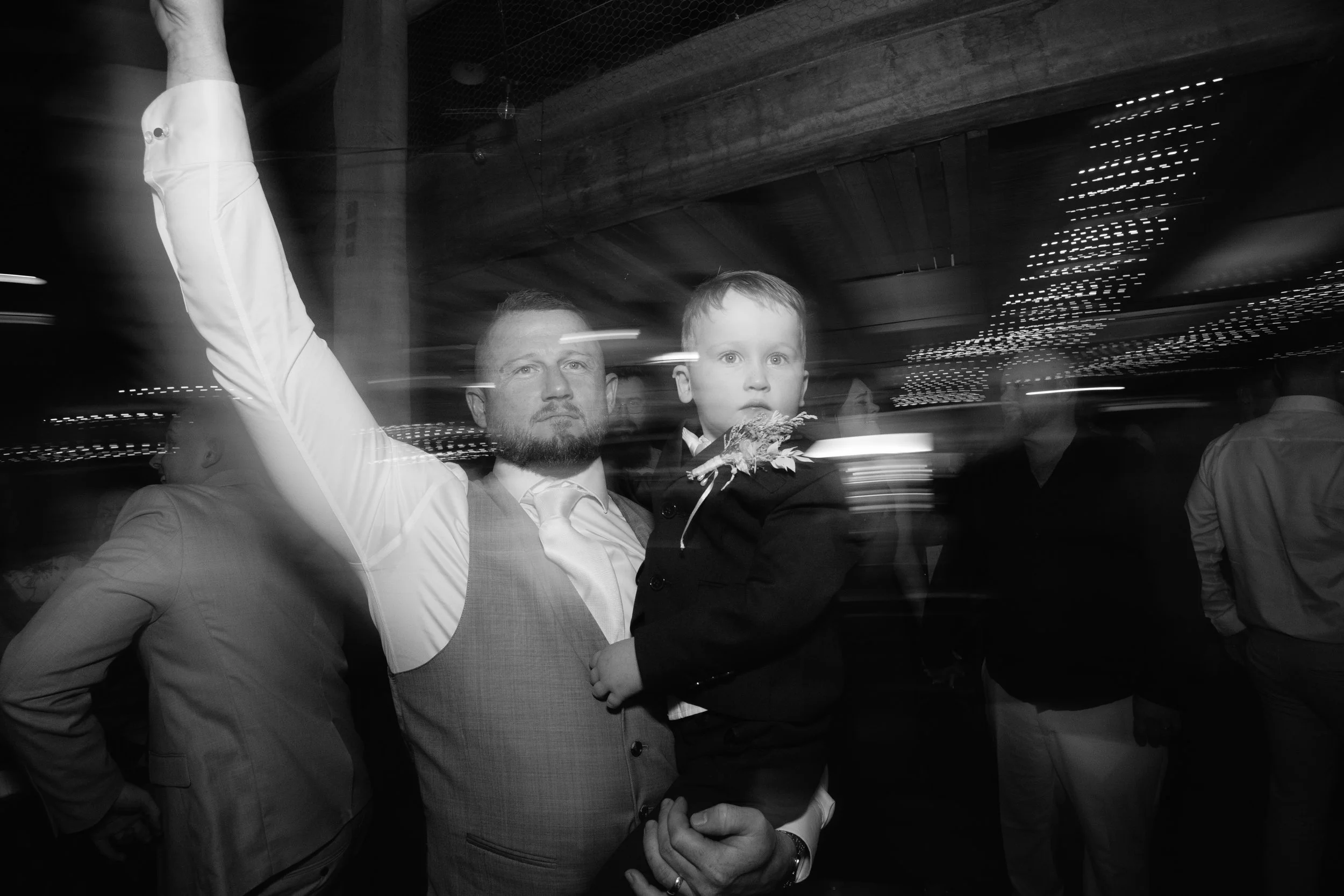 A man holding a young boy, both dressed in suits, at a social event in a dimly lit room with string lights, with other people partially visible in the background.