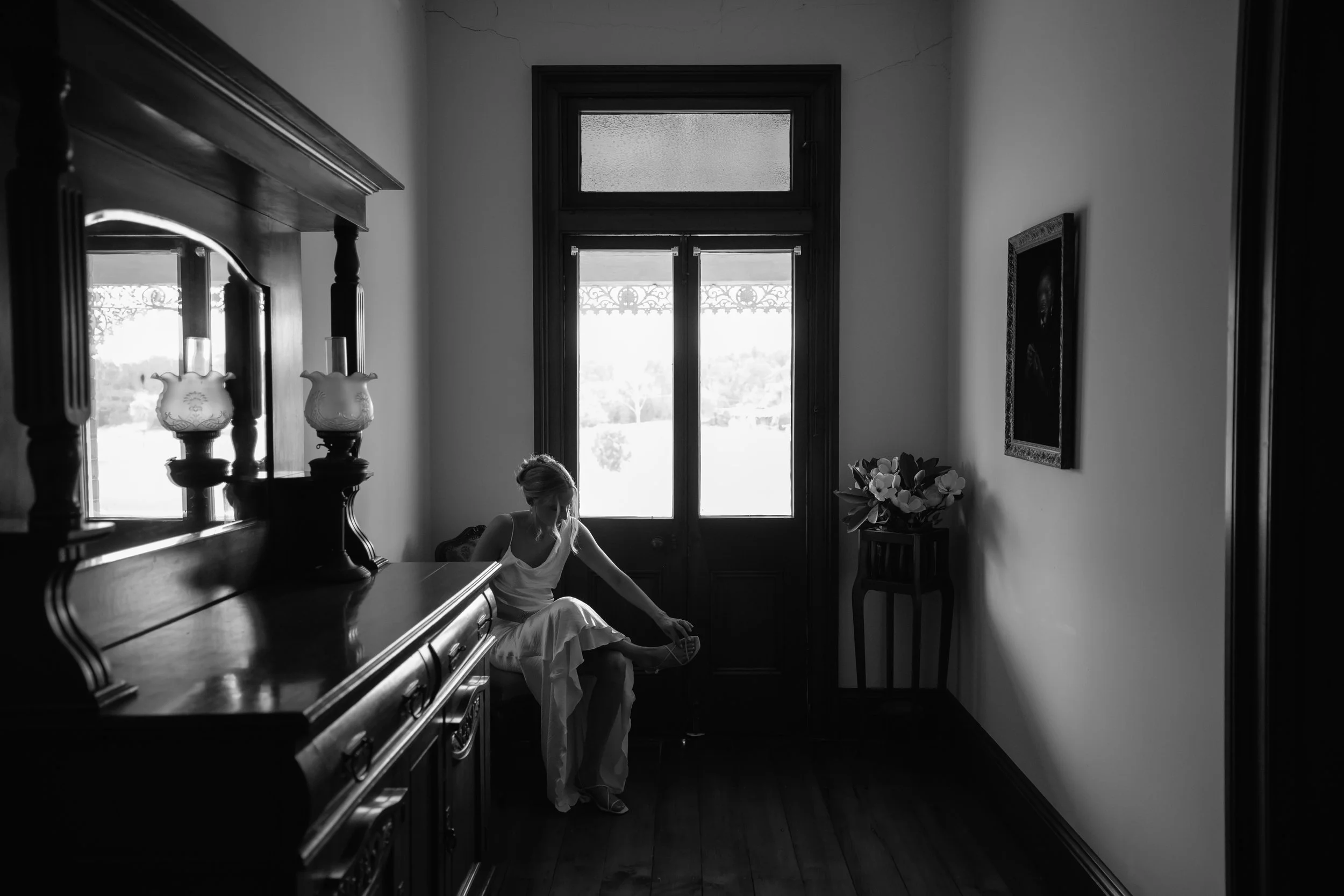 A black-and-white photograph of a woman sitting on a wooden floor, wearing a white dress, with her head bowed and hands on her lap. She is near a large window with decorative ironwork and a smaller window above it, in a vintage-style room with a vase