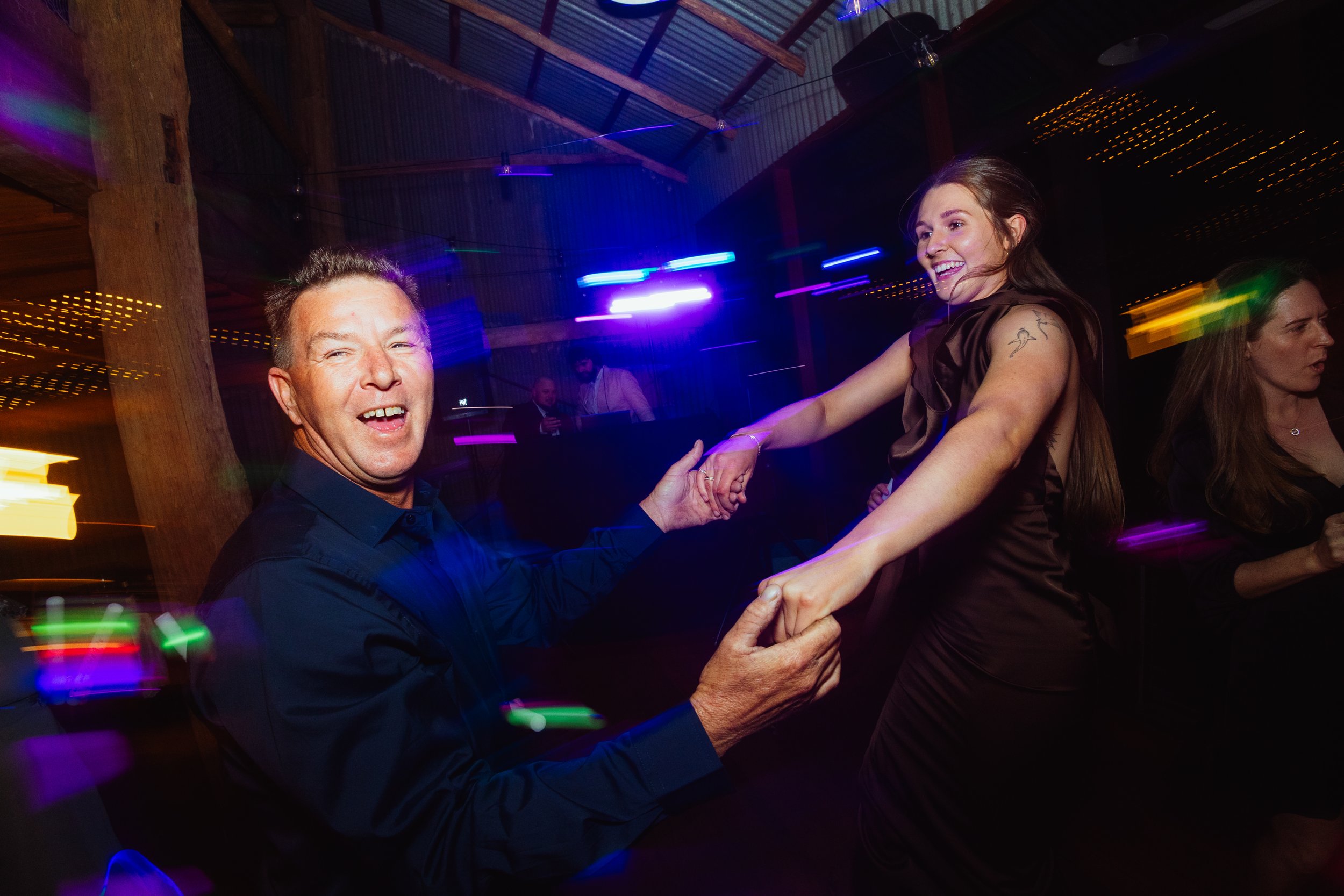 People dancing at a party with colorful lights and a rustic ceiling, smiling and holding hands.