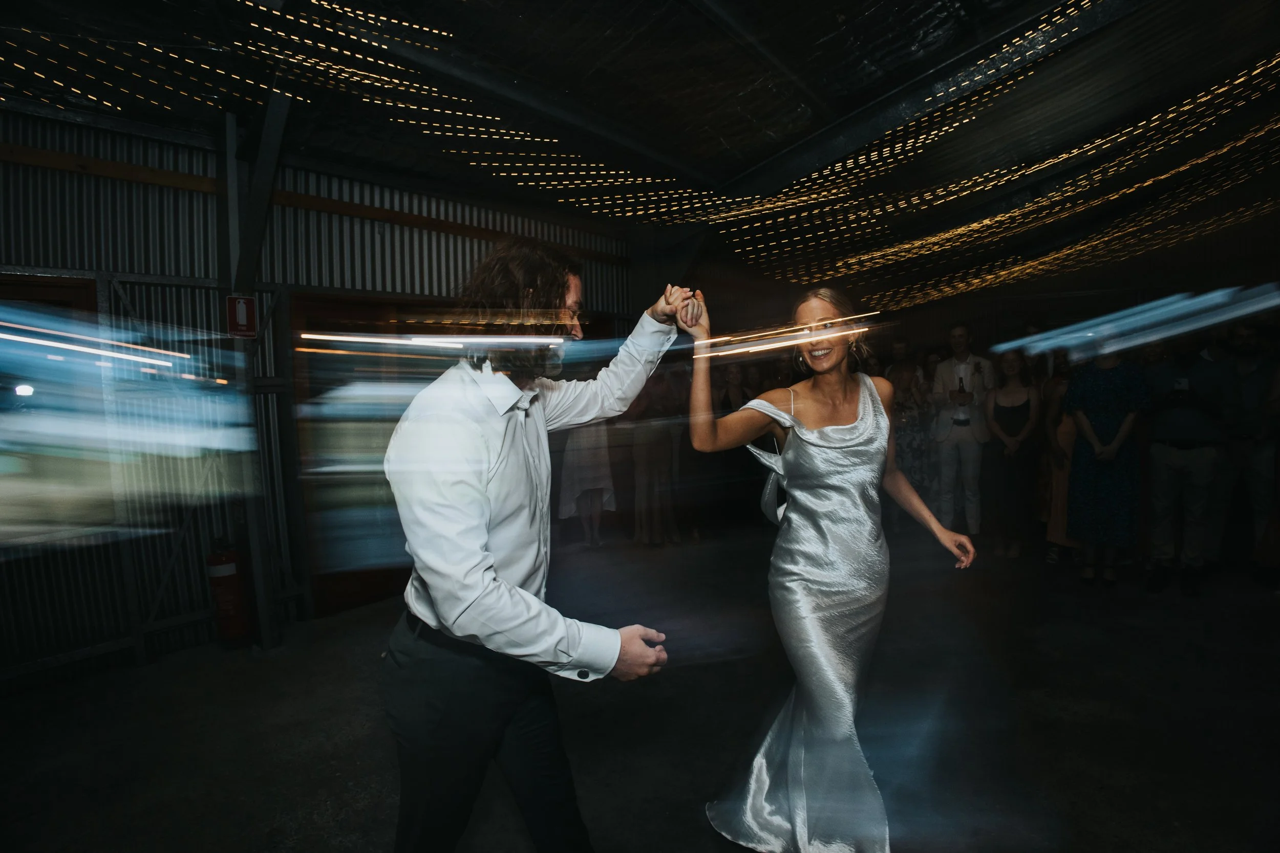 A man and woman dancing together at a celebration or wedding reception, with the man lifting the woman's hand as she twirls in her silver satin dress, surrounded by guests and string lights.