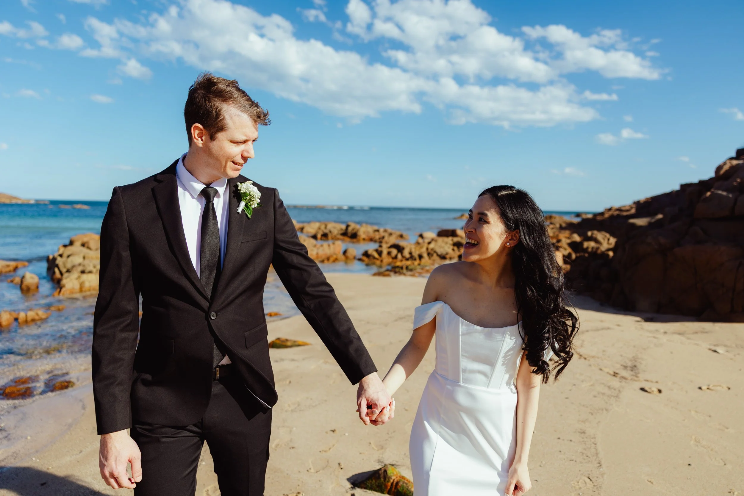 A newly married couple holding hands on the beach, with the groom in a black tuxedo and the bride in a white wedding dress, smiling at each other under a blue sky with scattered clouds.