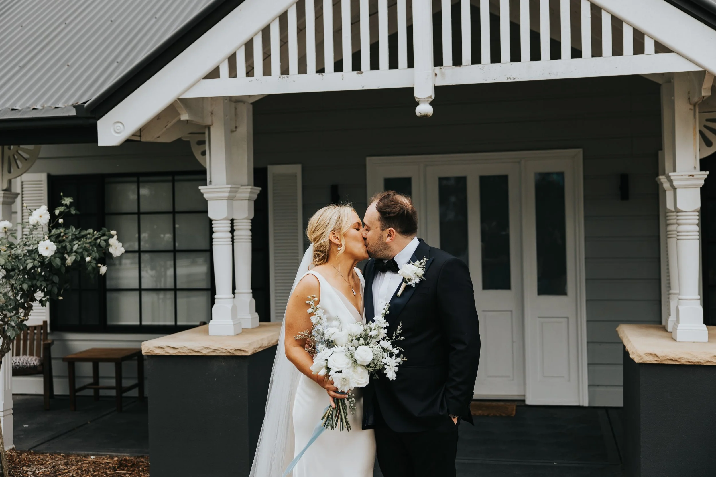 A bride and groom kissing in front of a white porch balcony at a wedding ceremony. The bride is holding a bouquet of white flowers, and the groom is dressed in a black suit and bow tie.