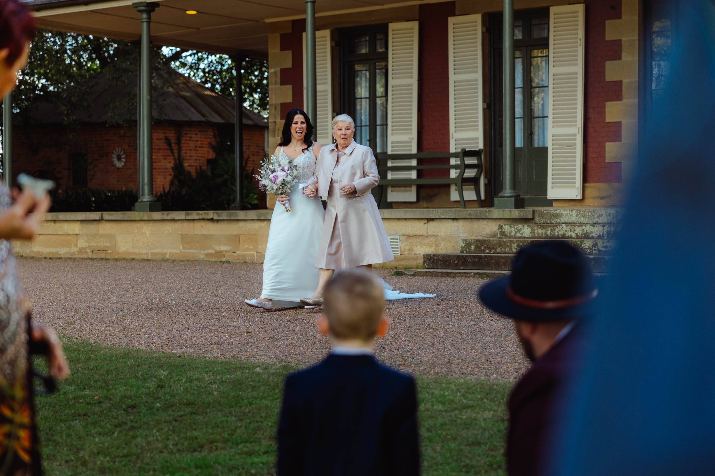 A wedding ceremony taking place outdoors in front of a brick house, with a bride in a white dress holding a bouquet and an elderly woman walking beside her, surrounded by seated guests.
