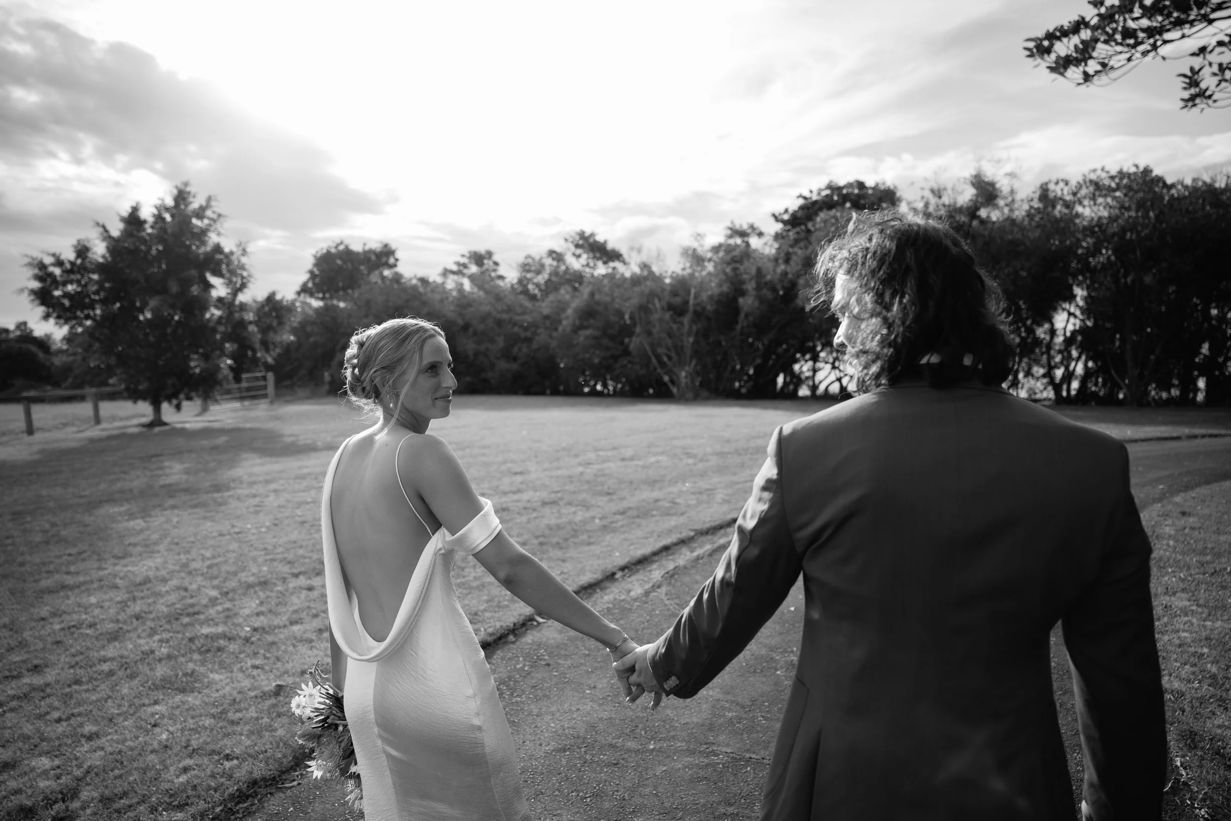 A black and white photo of a woman and man holding hands outdoors during sunset. The woman is wearing a white dress with thin straps, and the man is dressed in a suit. They are looking at each other, with trees and a cloudy sky in the background.